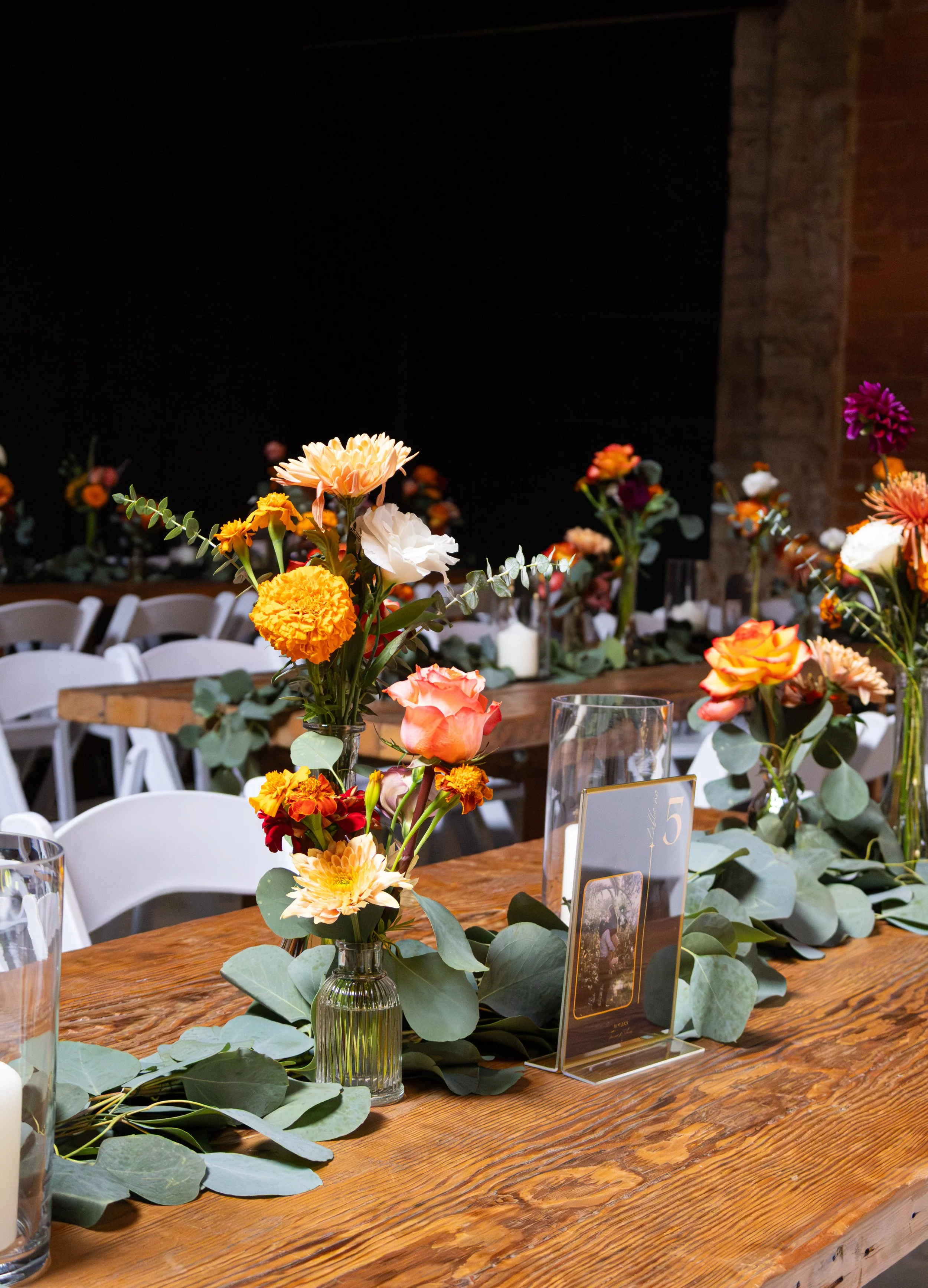 Flower table centerpiece with orange, white, and pink flowers, green leaves, and a small table card with number 5, on a rustic wooden table setting at event.