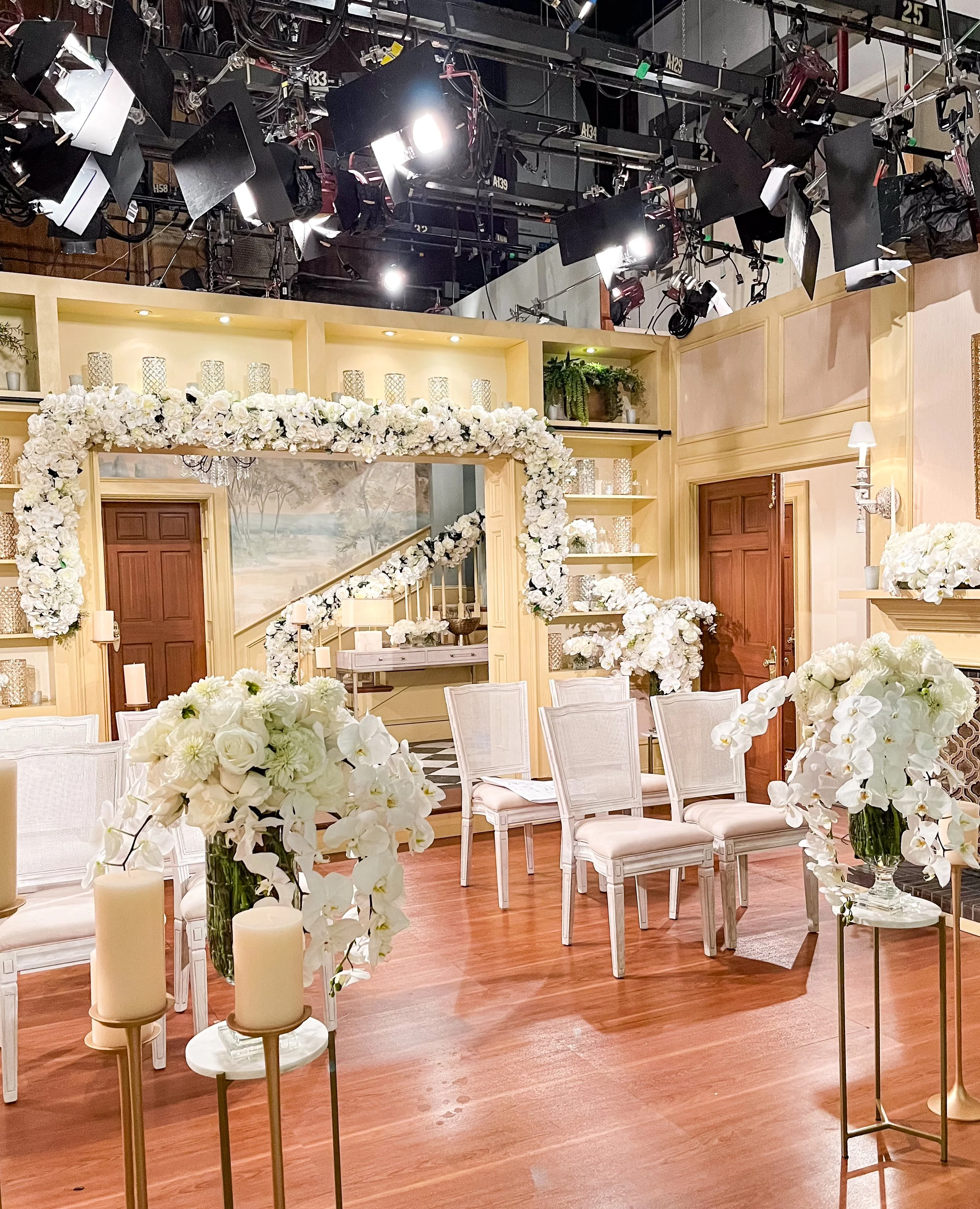A decorated indoor wedding ceremony setup with white floral arrangements, candles, chairs, and a floral arch, on a wooden floor, with lighting and film equipment overhead.