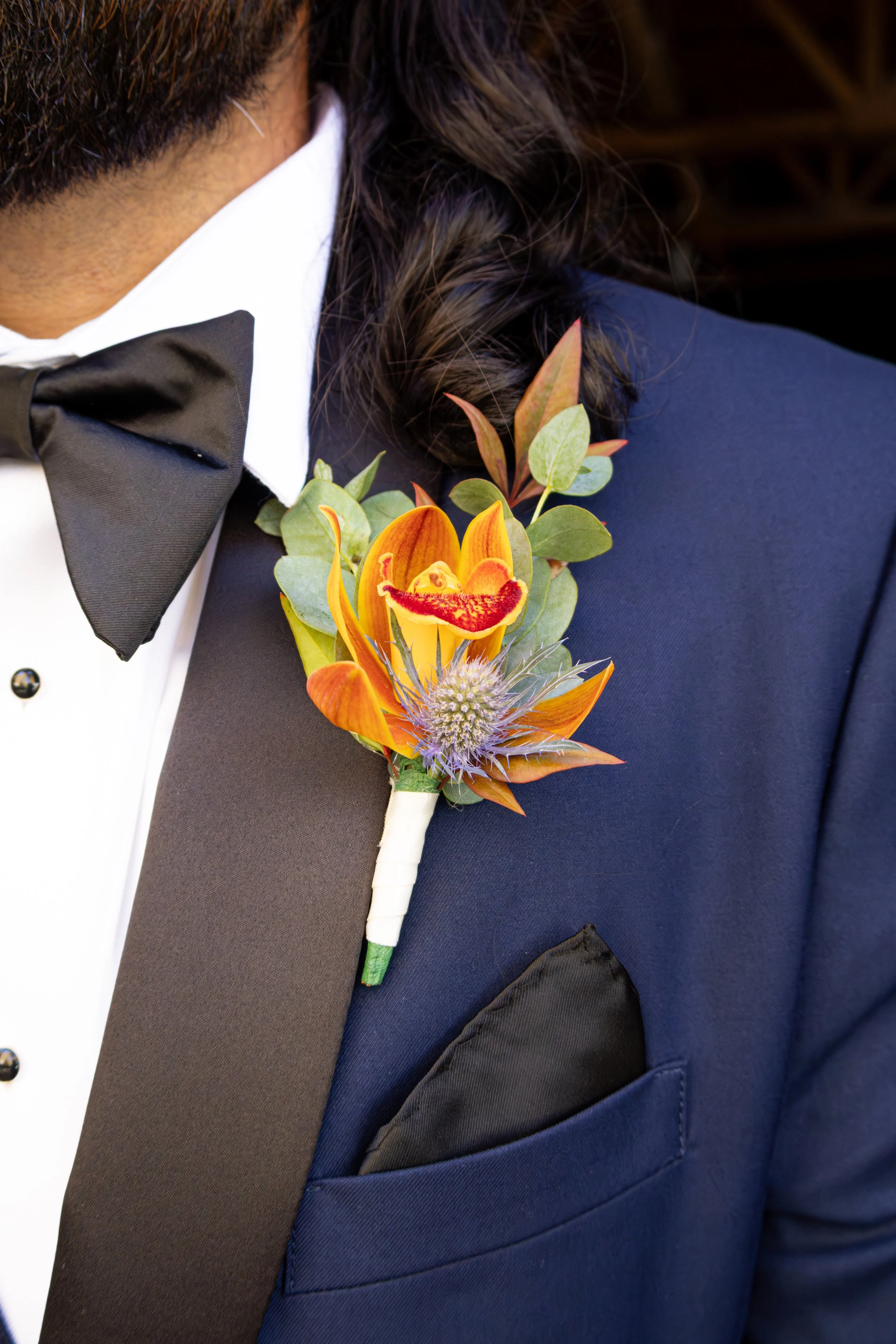 Close-up of a man in a navy blue tuxedo jacket with a black satin lapel, white shirt, and black bow tie, wearing a colorful floral boutonniere on his lapel.