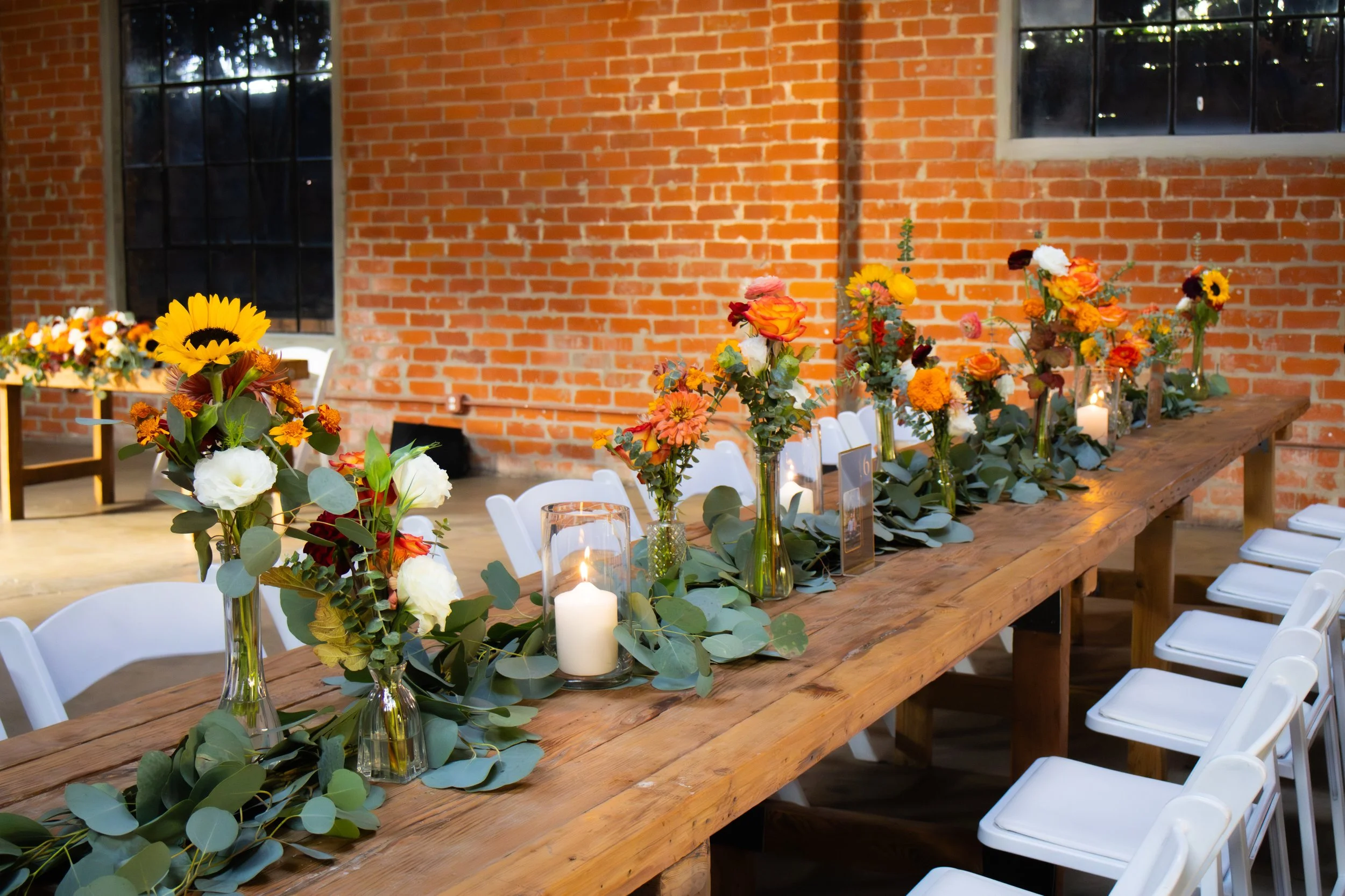 Long wooden table decorated with colorful flower arrangements, candles, and greenery, set up for a celebration in a brick-walled venue with large windows.