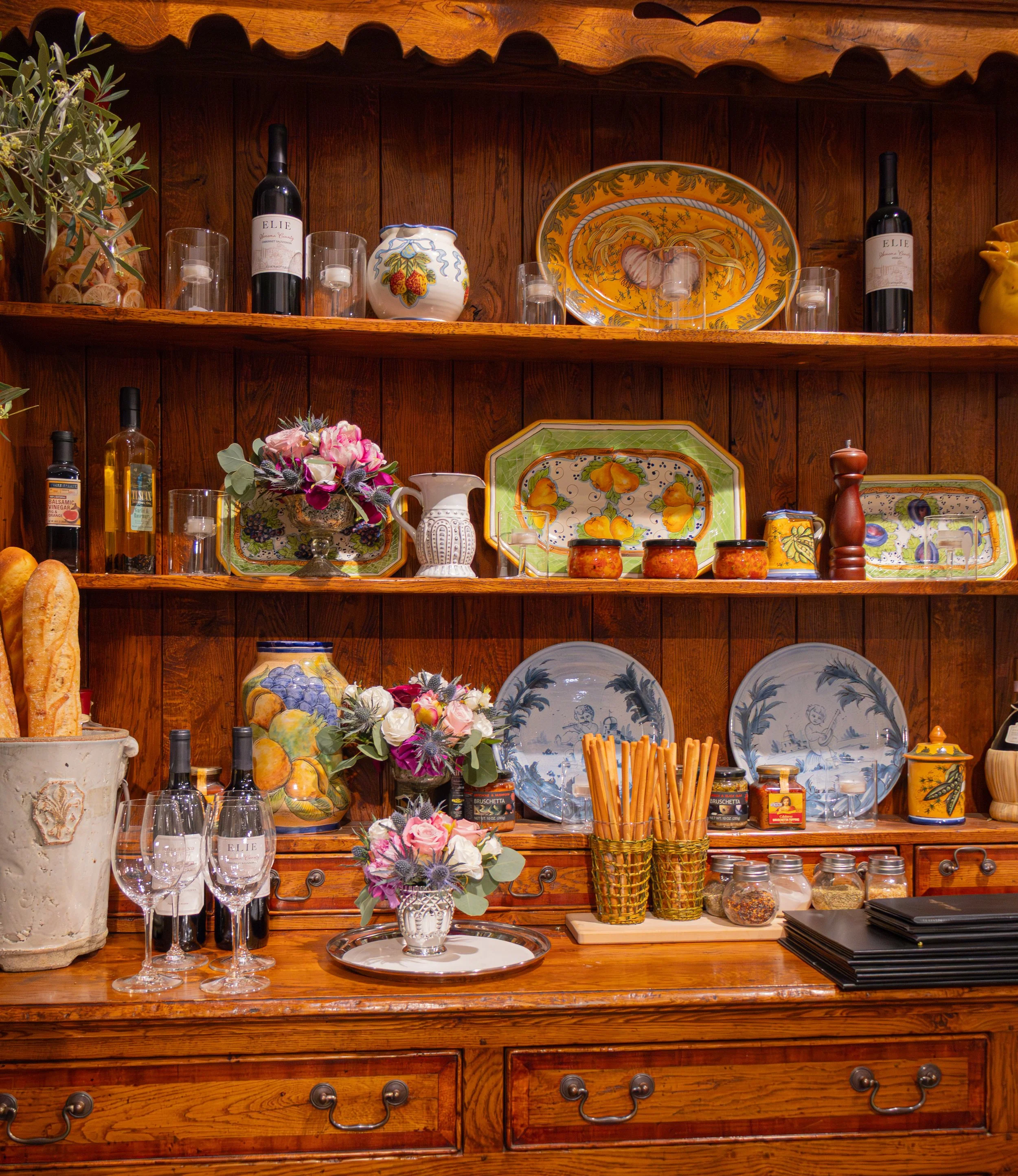A wooden kitchen hutch with shelves displaying decorative plates, flowers in vases, wine bottles, glassware, and jars of spices and preserves. The top shelf has wine bottles, glasses, and a decorative plate. The middle shelf features colorful ceramic