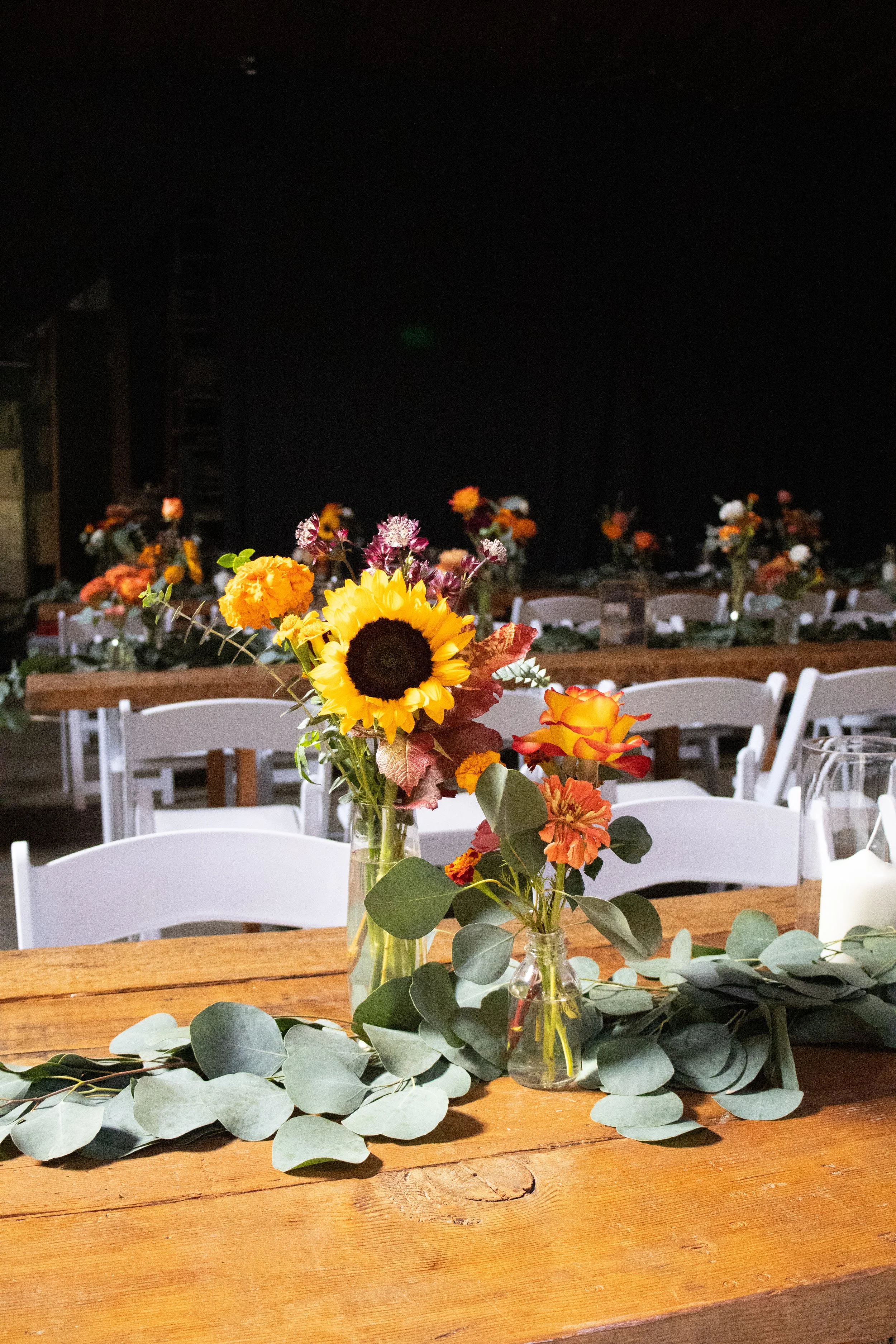 A wooden table decorated with a green eucalyptus garland, featuring two small glass vases with colorful flowers, including sunflowers, roses, and daisies, set in a dimly lit event space with additional floral arrangements in the background.