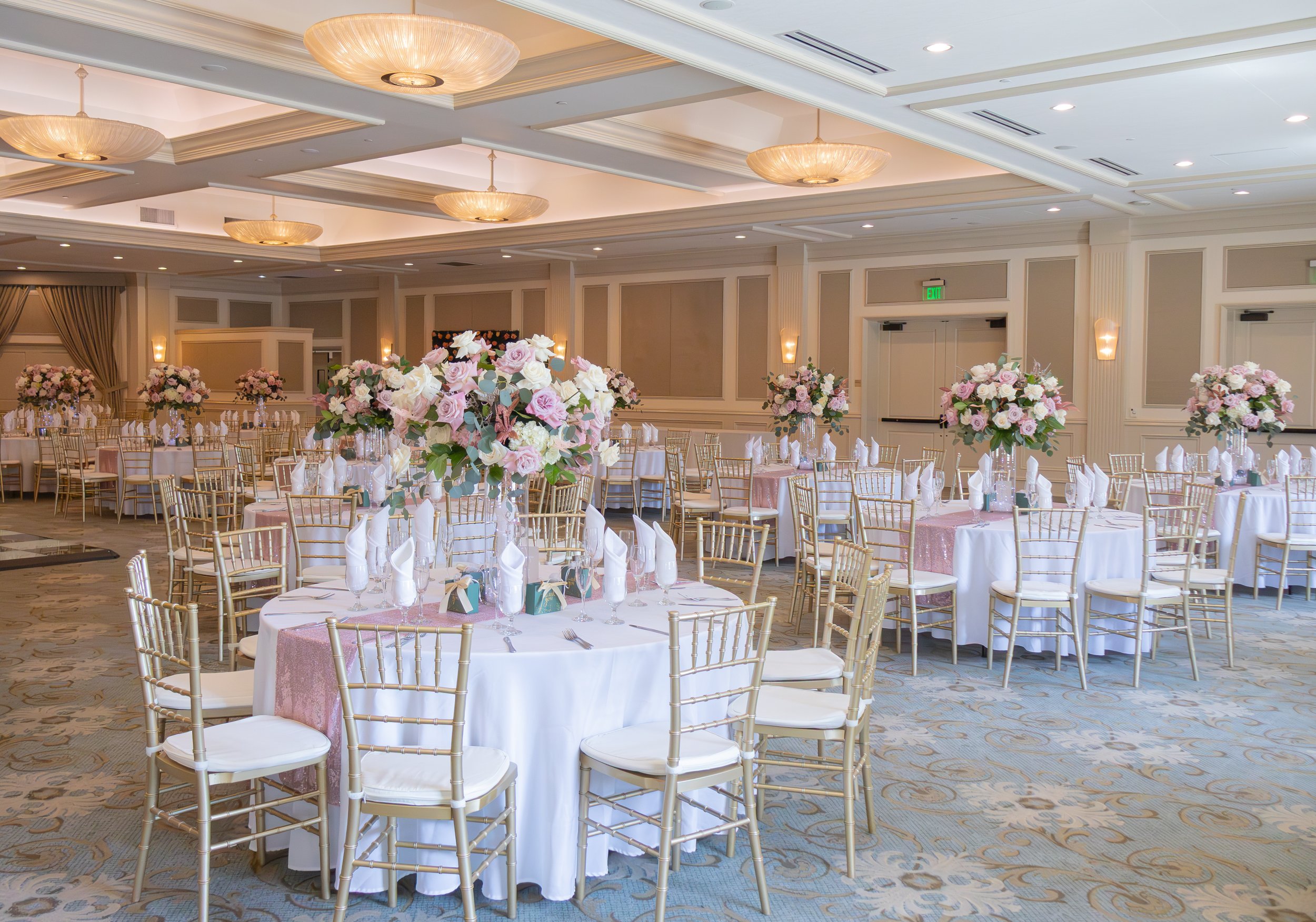 Elegant banquet hall decorated for a wedding reception, with round tables covered in white tablecloths and tall floral centerpieces with pink and white flowers, gold chairs, and soft lighting.