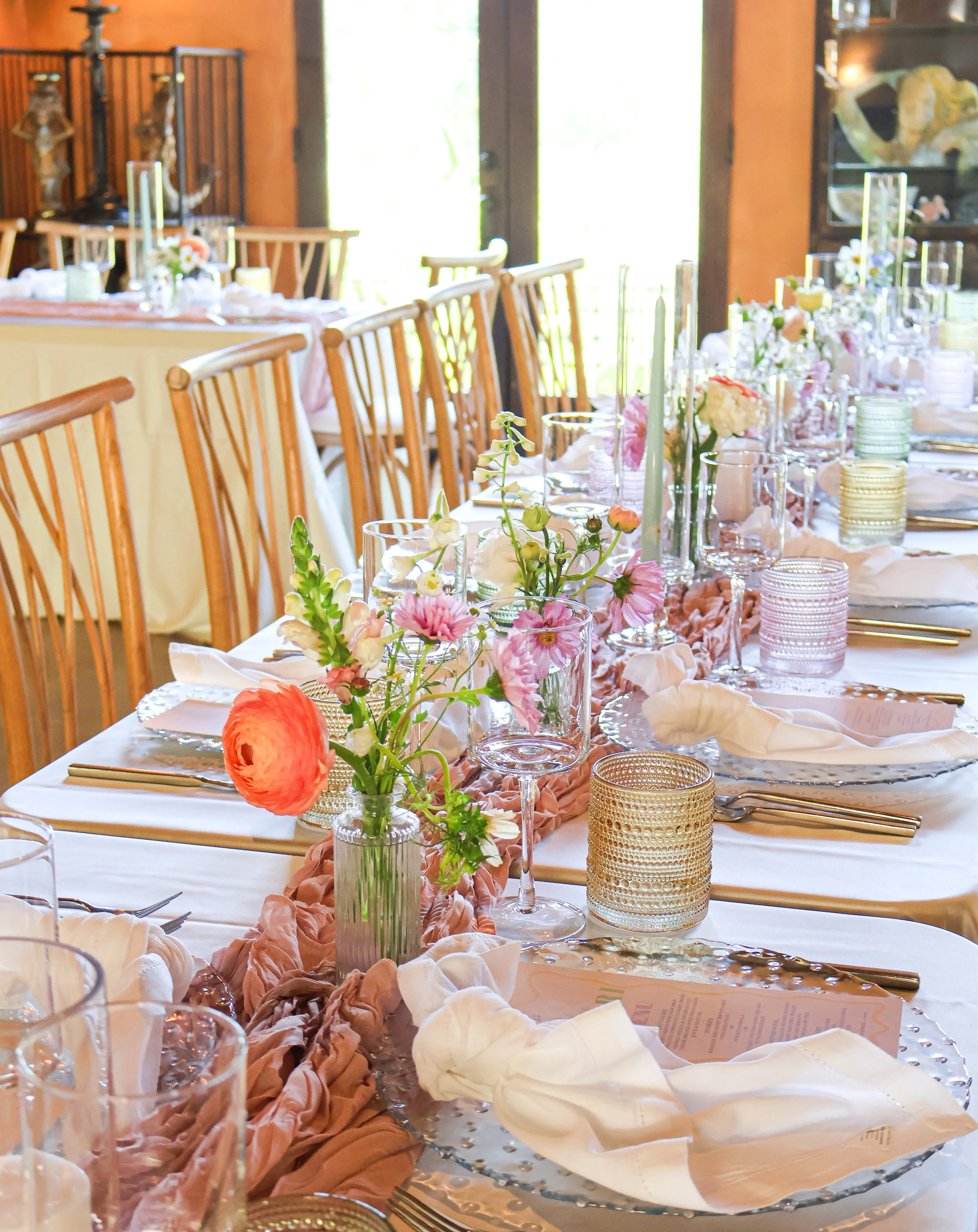 Elegant banquet table set with floral centerpieces, candles, glassware, and gold-colored flatware in a sunny, warmly decorated room.