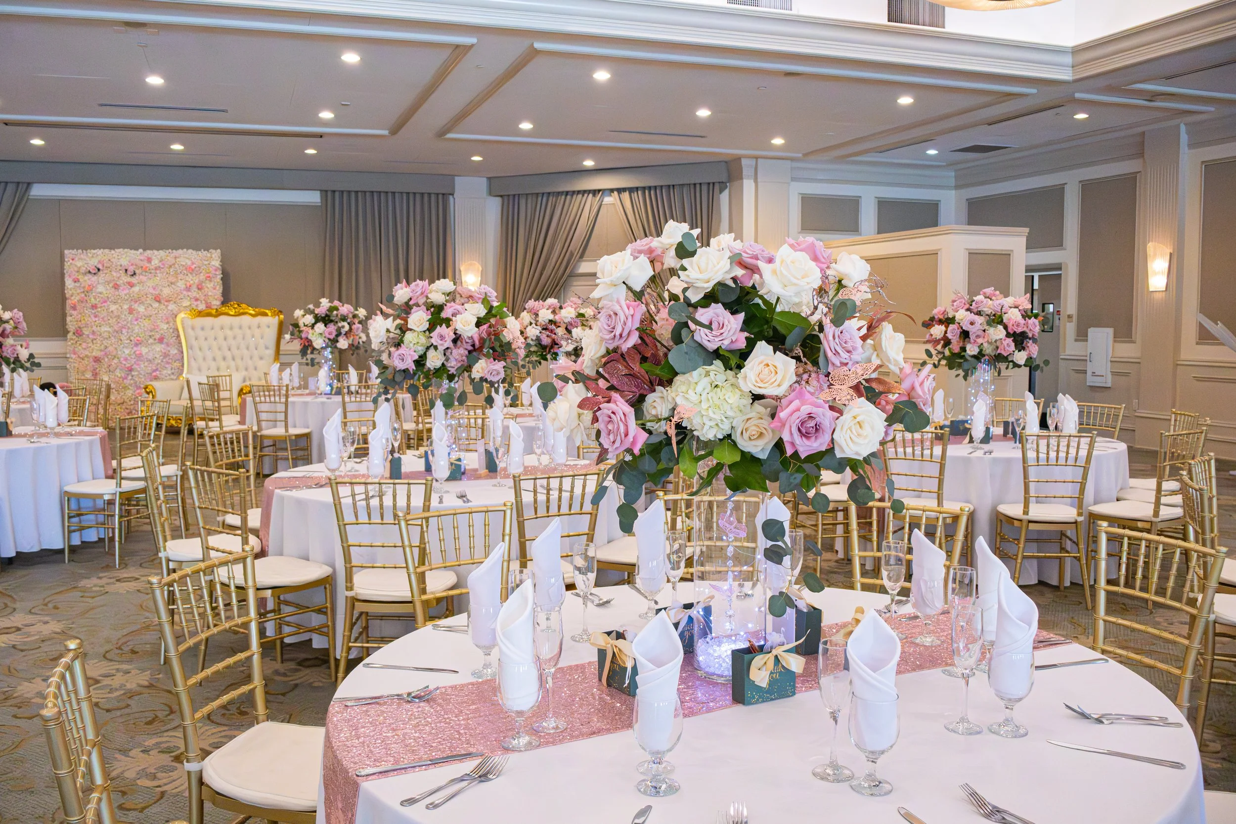 Elegant banquet hall decorated for a wedding or special event, featuring round tables with pink tablecloth runners, tall floral centerpieces with pink and white roses, gold chairs, and neatly folded white napkins.