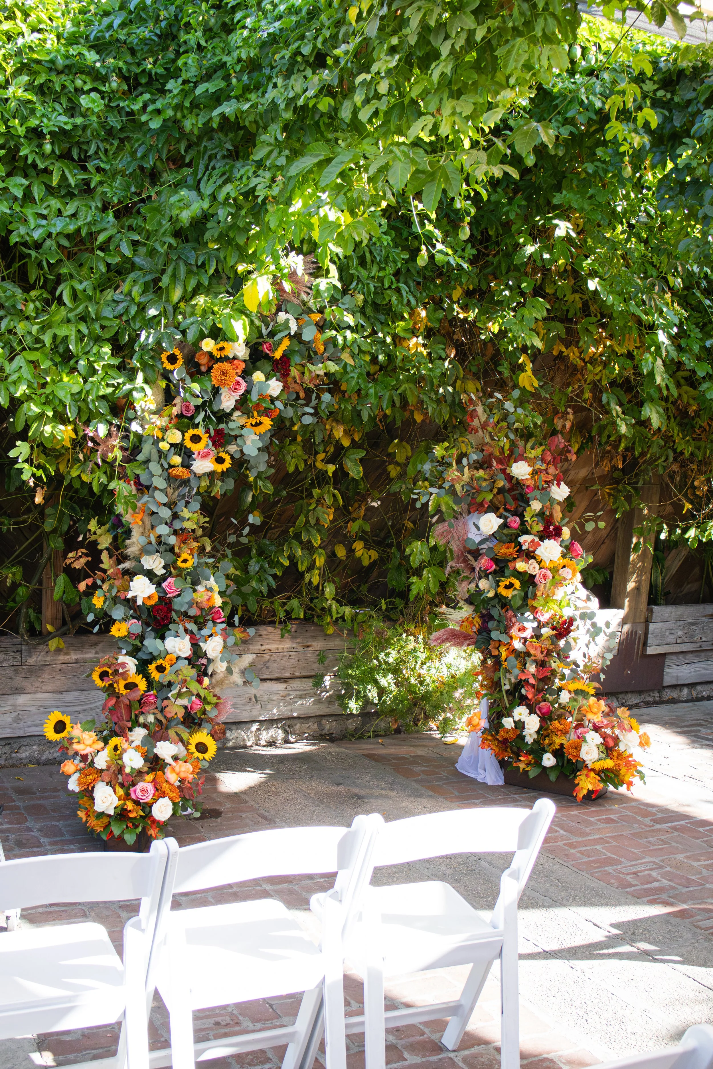 A floral arch decorated with colorful flowers including sunflowers, roses, and other blooms, set against a lush green leafy background, with white chairs in the foreground, possibly for a wedding or outdoor event.