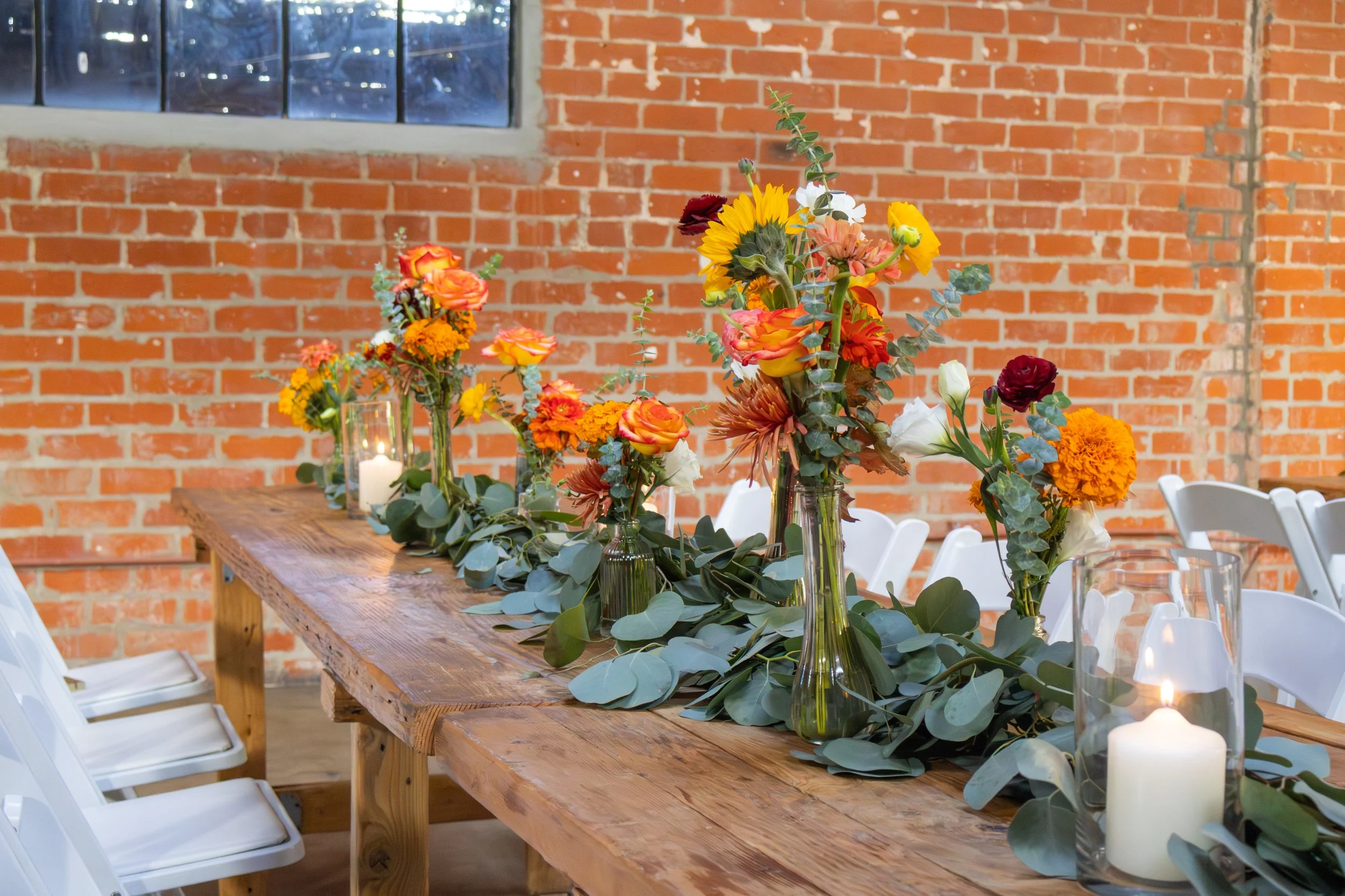 Long wooden table decorated with colorful flower arrangements in vases, surrounded by white chairs, against an exposed brick wall with a window in the background, and candles in glass placements along the centerpiece.