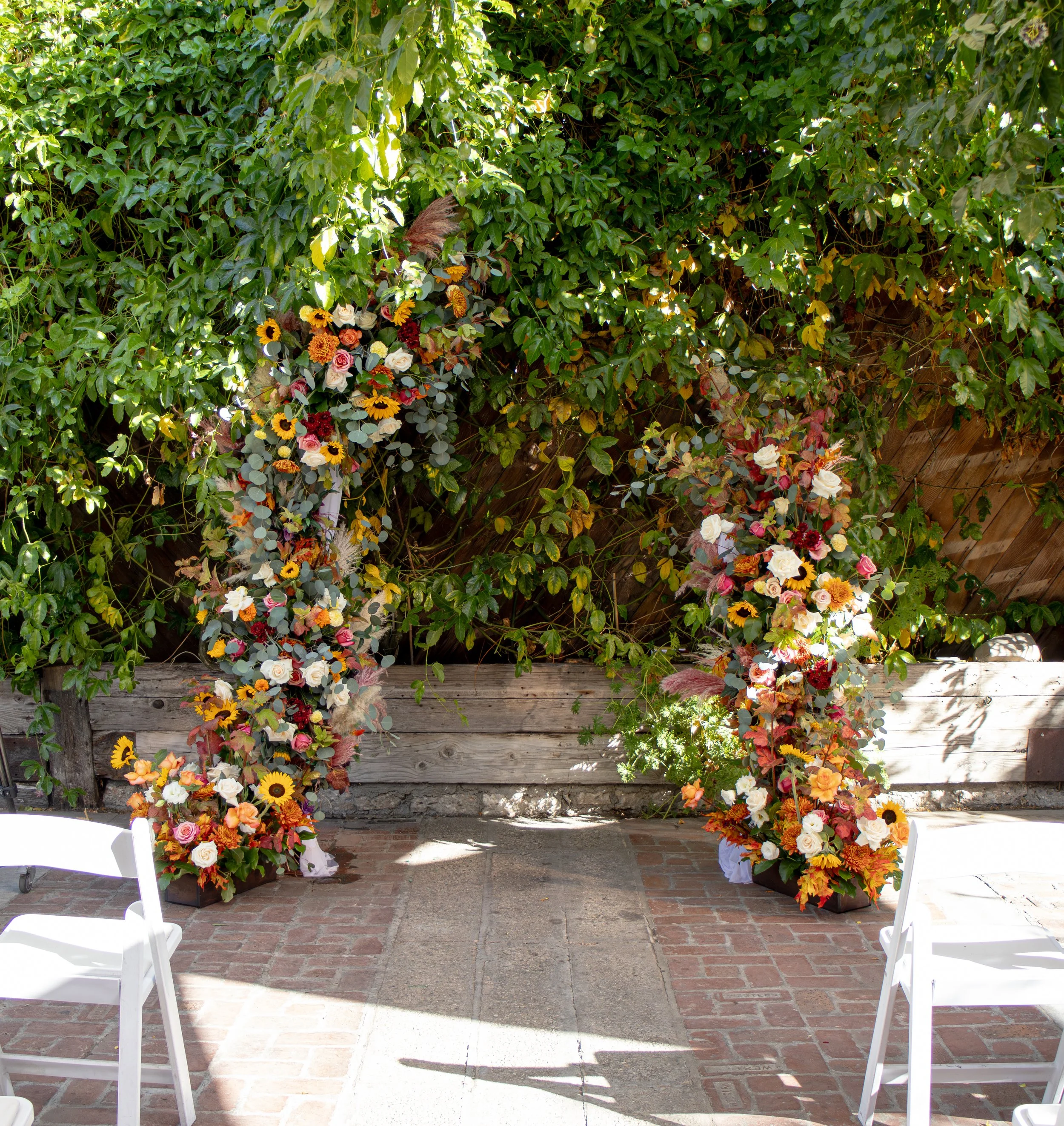 Decorative floral arch with colorful flowers and greenery set up outdoors on a brick patio, with white chairs nearby.