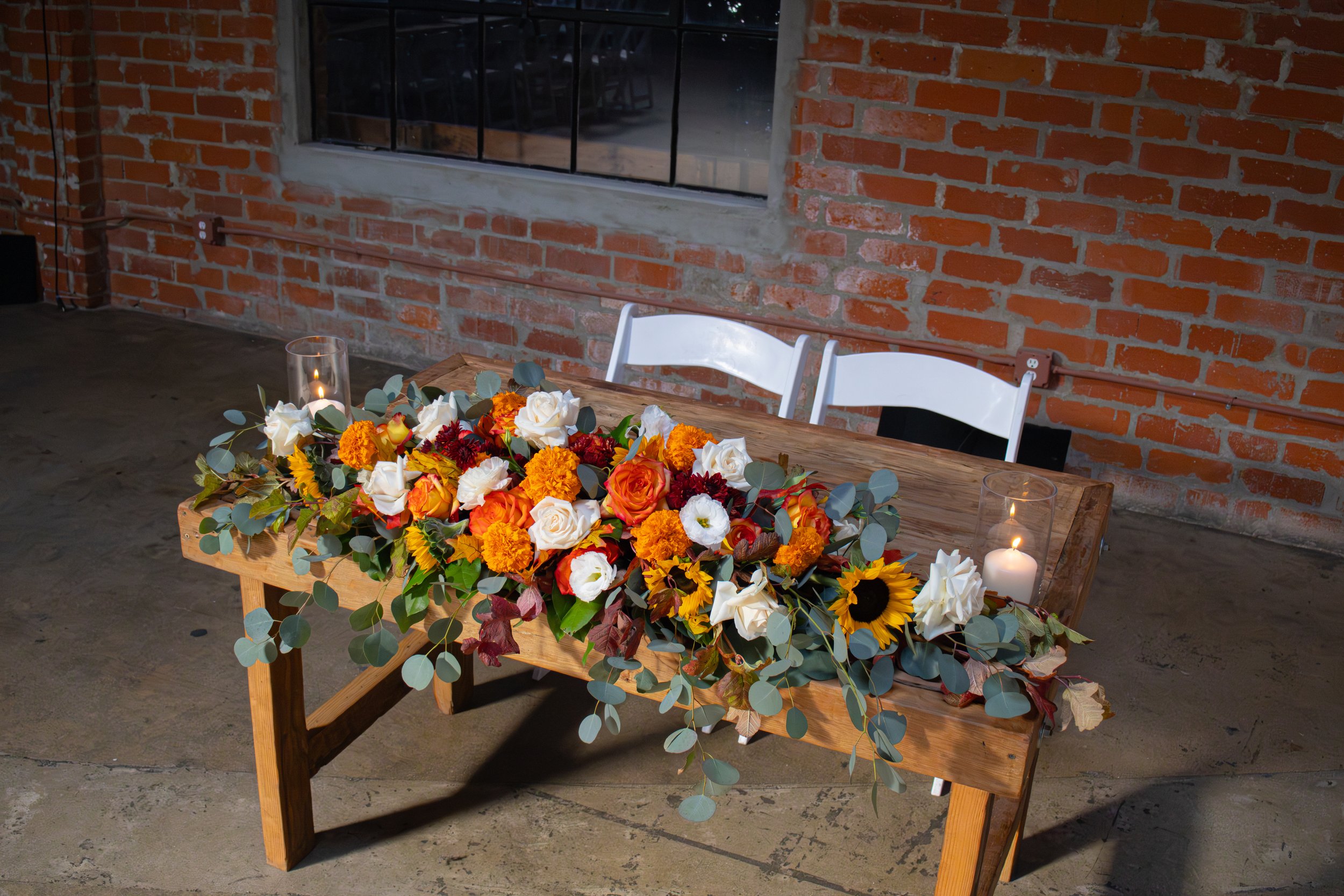 A wooden table decorated with a large colorful flower arrangement and two glass candle holders with lit white candles. The background features a brick wall and a large window.