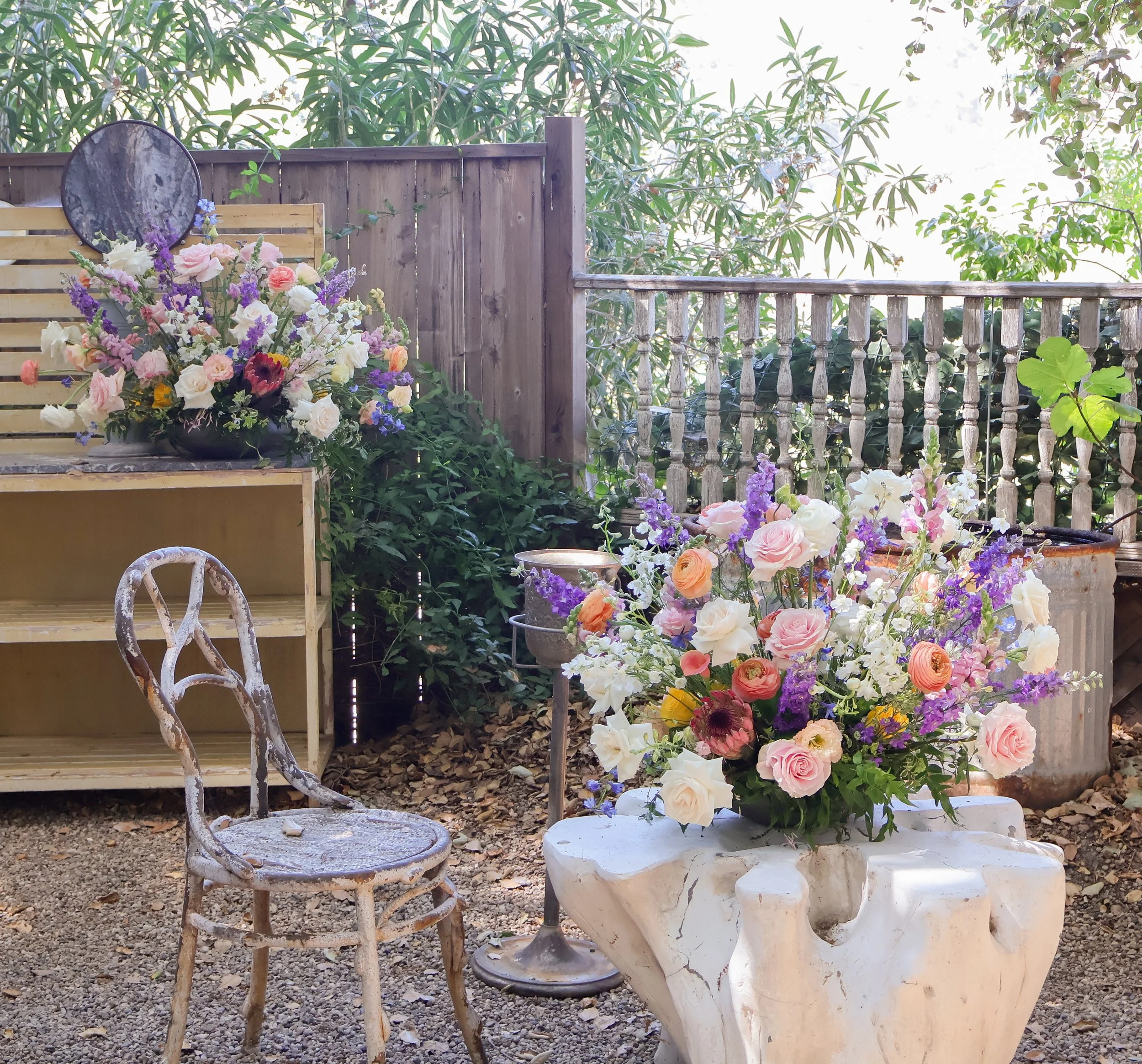 Outdoor garden scene with large colorful flower arrangements on a white stone table and a rusted metal chair, wooden shelves, potted plants, and a wooden fence.