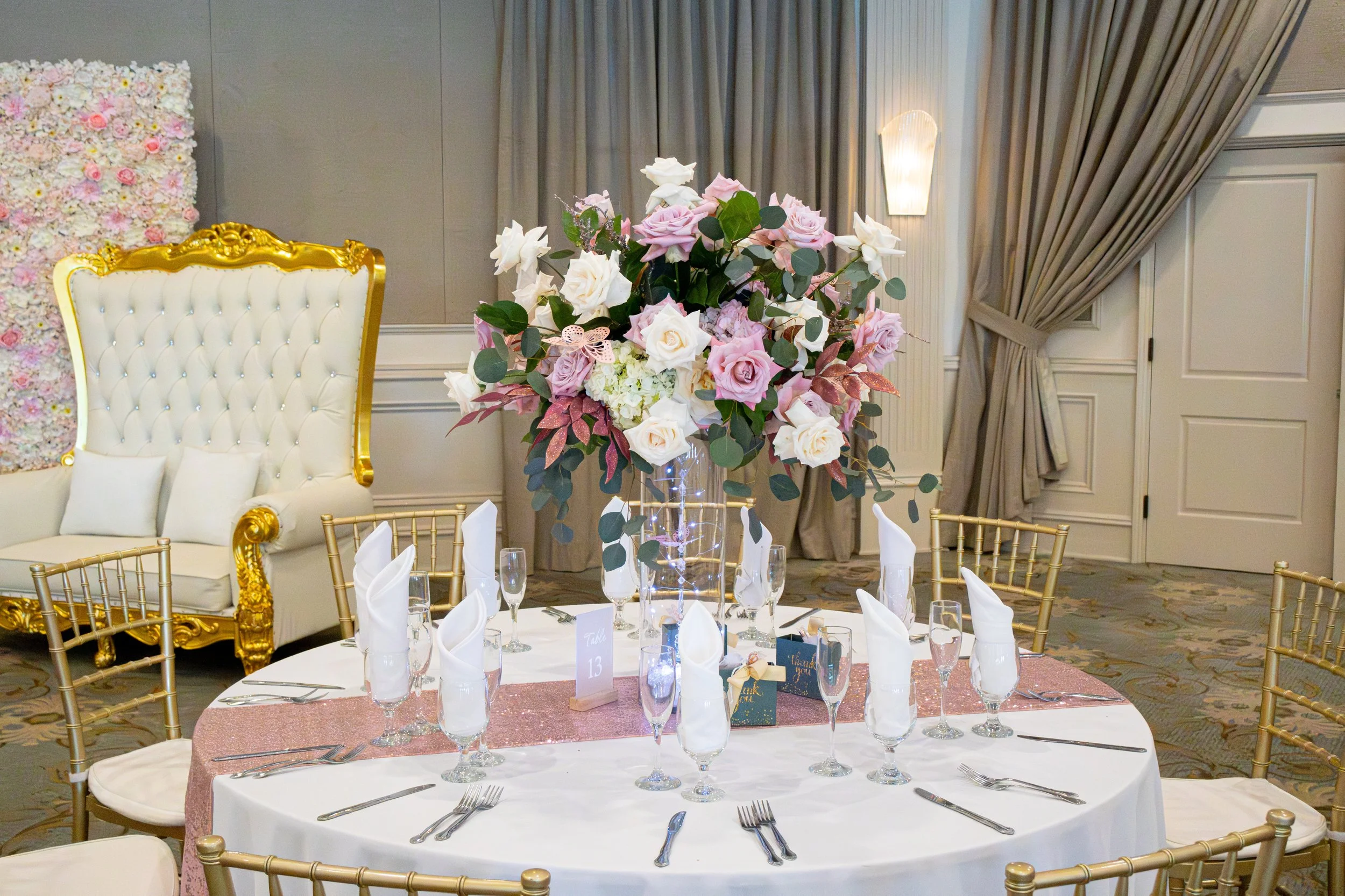 A decorated wedding reception table with a floral centerpiece of pink and white roses, surrounded by gold chairs in a banquet hall with beige drapes and wall lighting.