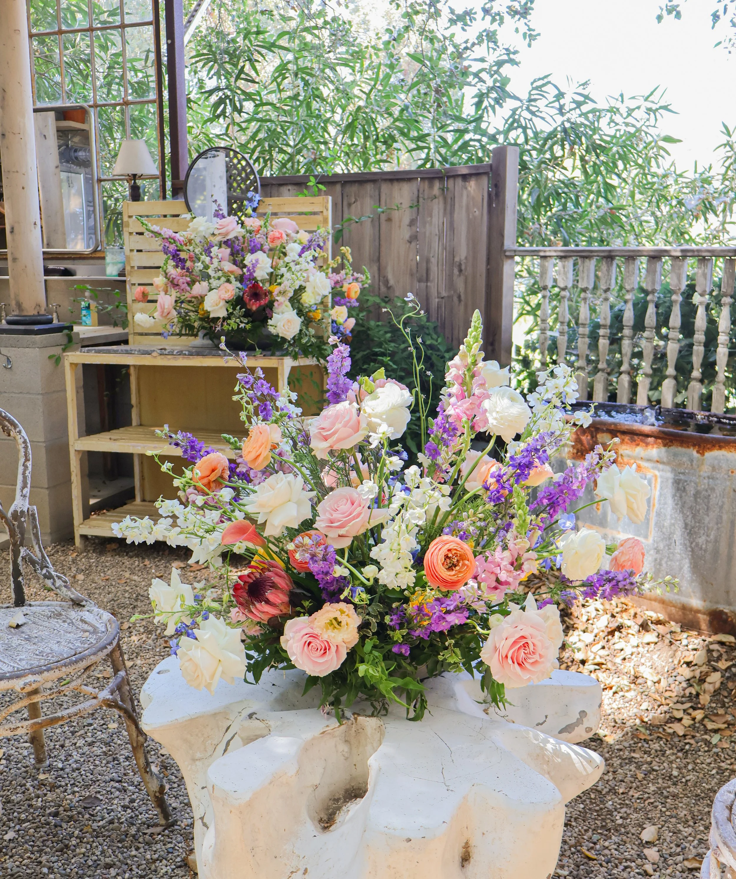 A large colorful flower arrangement with pink roses, purple, white and orange flowers, placed on a white sculptural table in an outdoor garden setting.