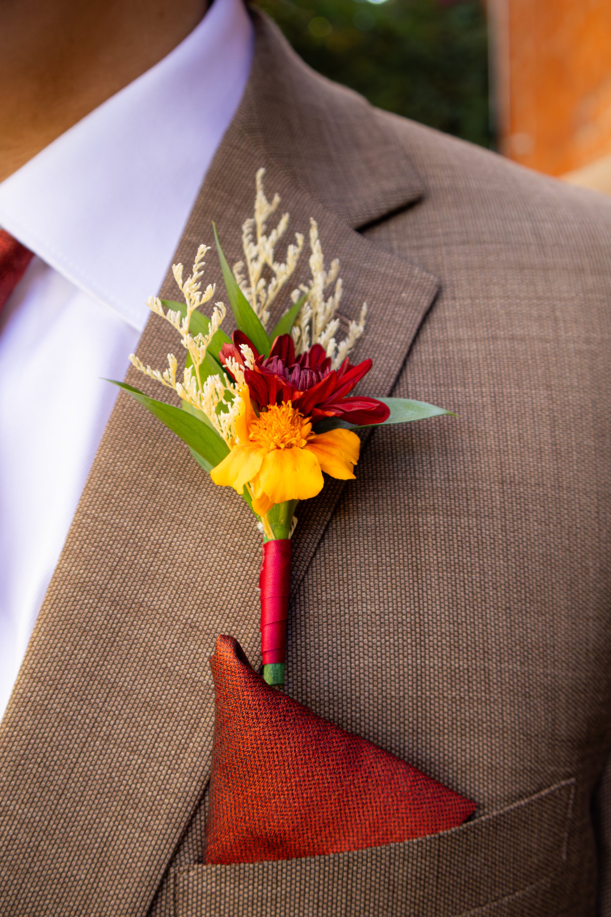 Close-up of a men's brown checkered suit with a white shirt, red tie, and a boutonniere featuring yellow, red, and purple flowers with green leaves, attached to the jacket's lapel.