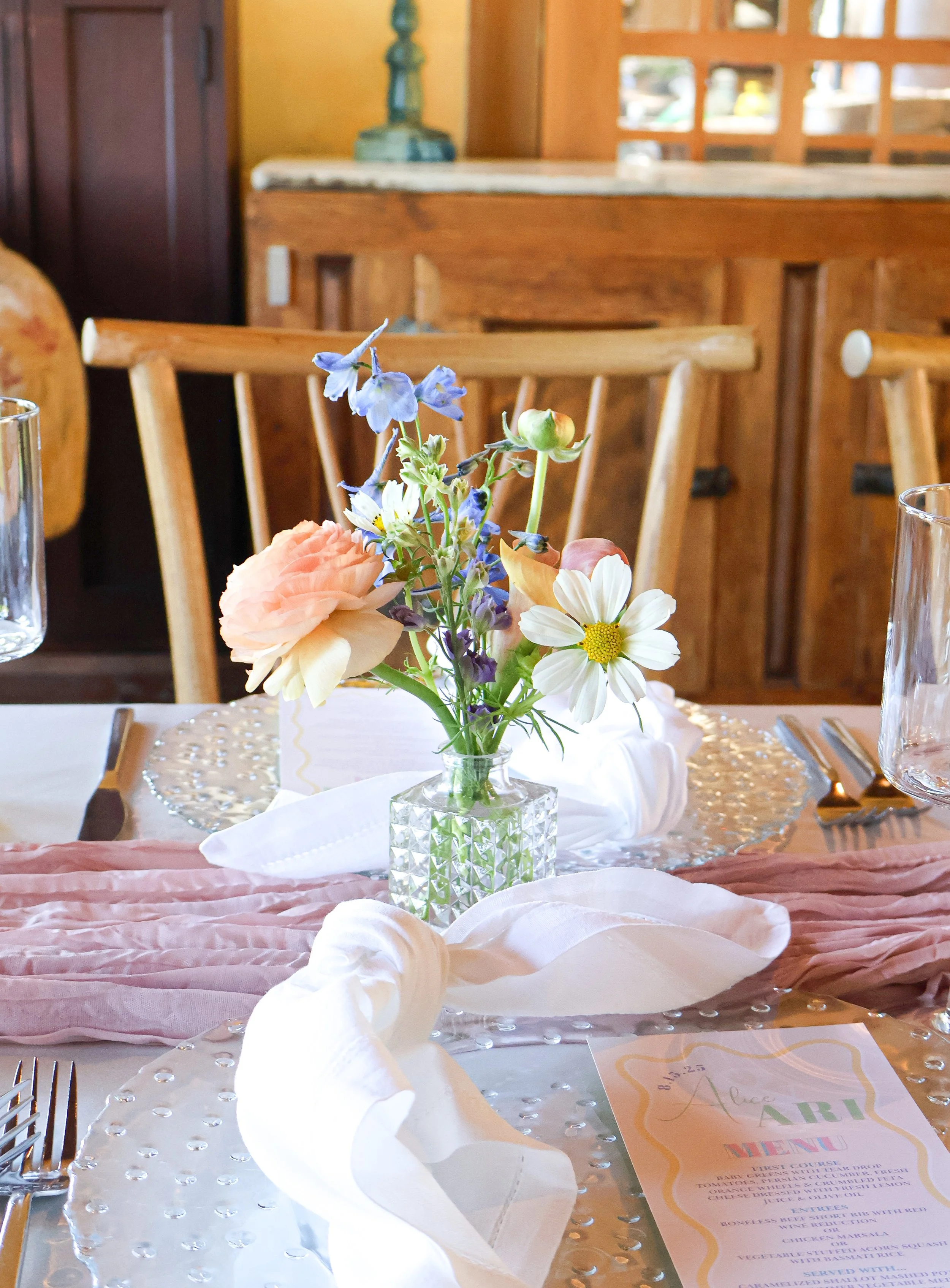 A floral centerpiece with pink roses, white daisies, and blue flowers in a clear textured glass vase on a table decorated for a special event.