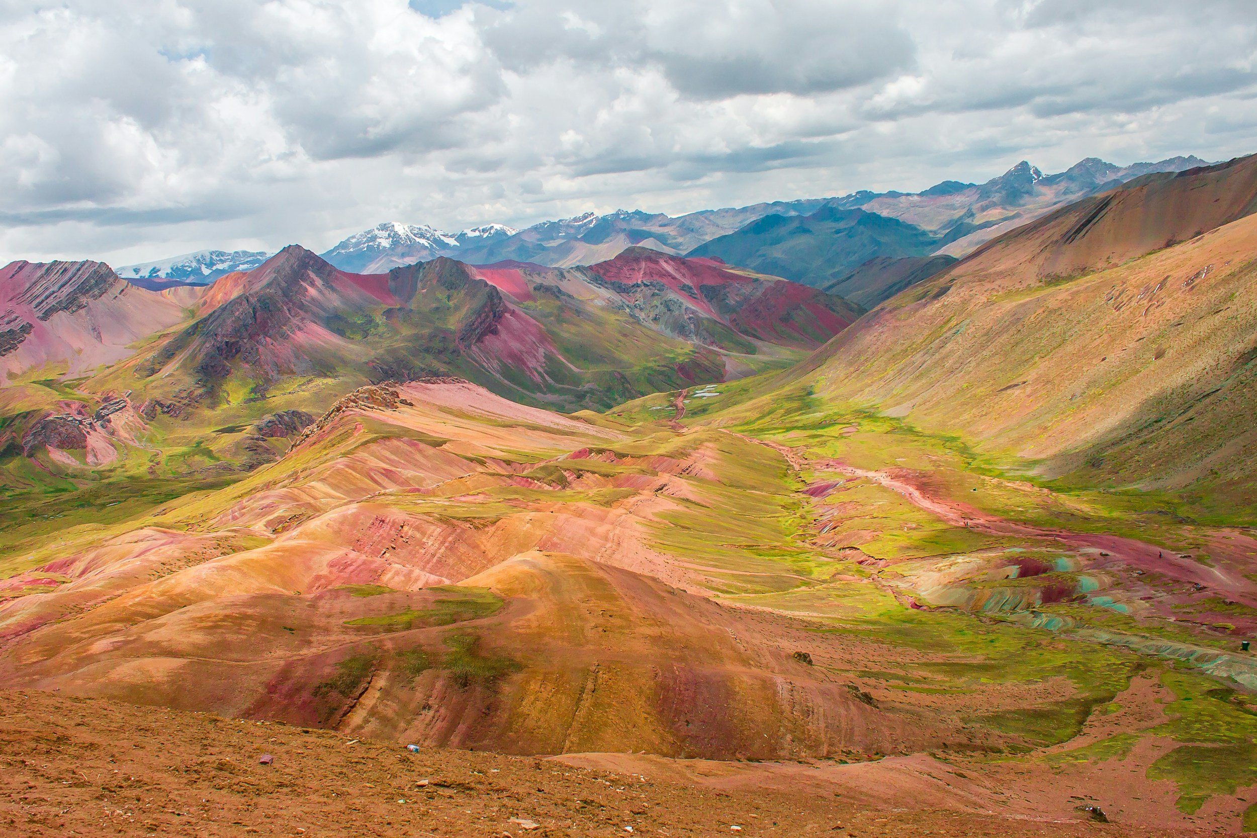 Colorful mountainous landscape with multi-colored hills, valleys, and distant snow-capped mountains under cloudy sky.