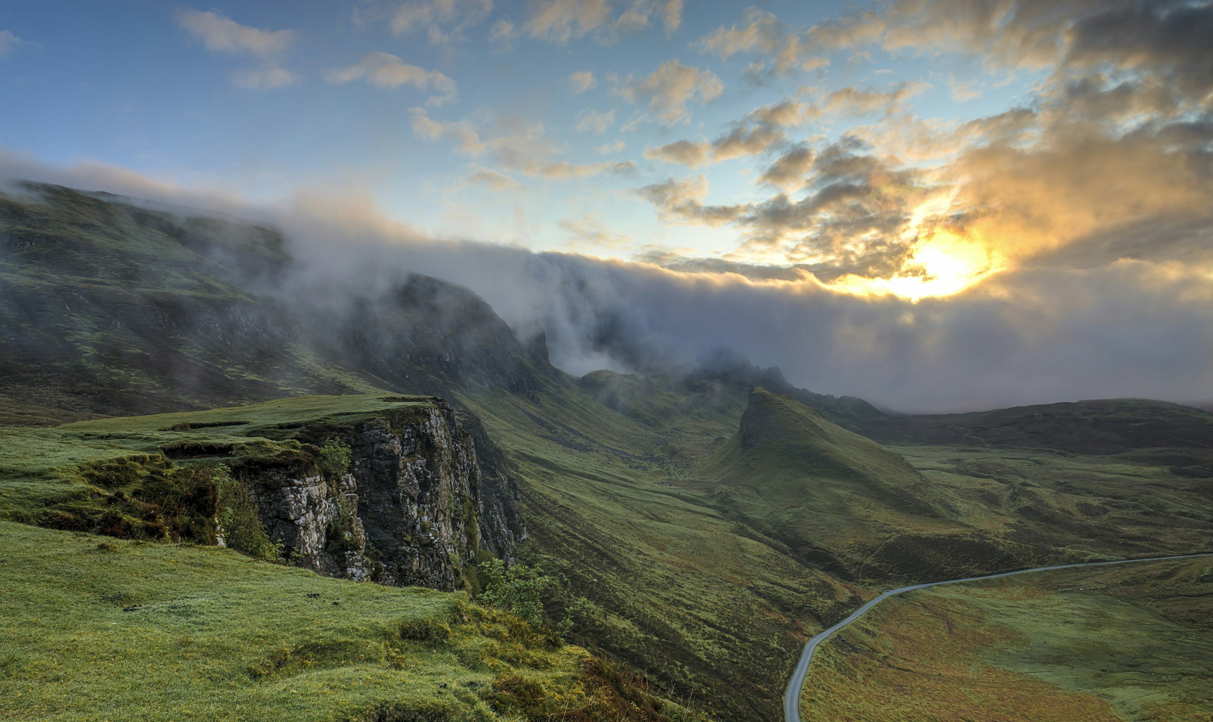 Scenic view of green rolling hills and cliffs with mist and clouds at sunset, a winding road in the valley.