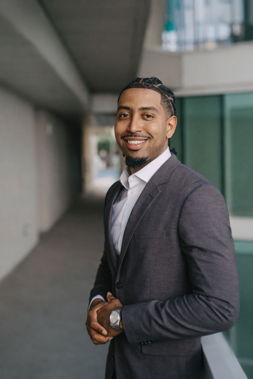 A smiling man in a gray suit and white shirt stands outdoors on a balcony, with modern buildings and a glass railing visible in the background.