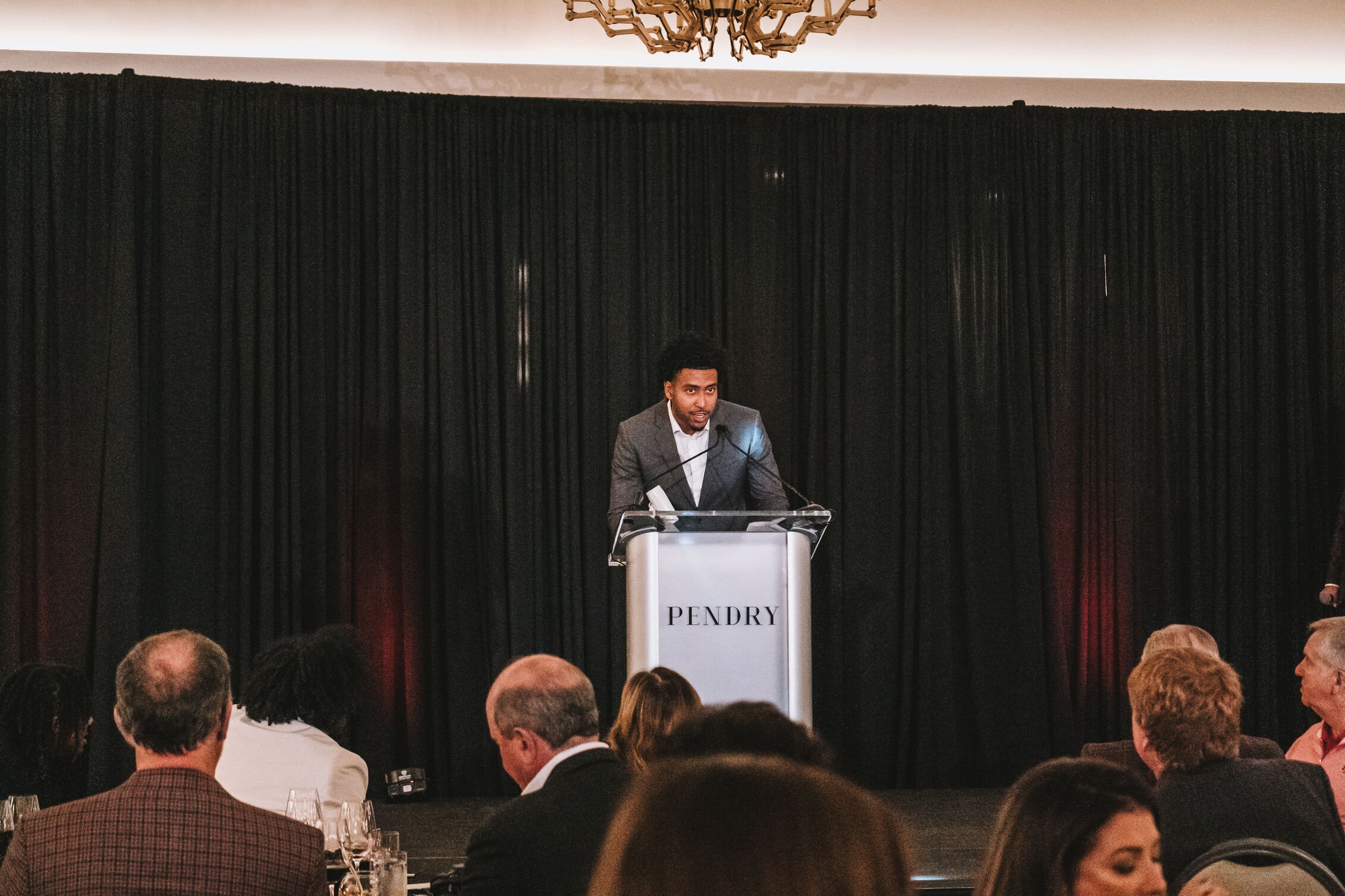 A man in a gray suit speaking at a podium with a microphone, in front of a black curtain background, at an event with seated audience members.