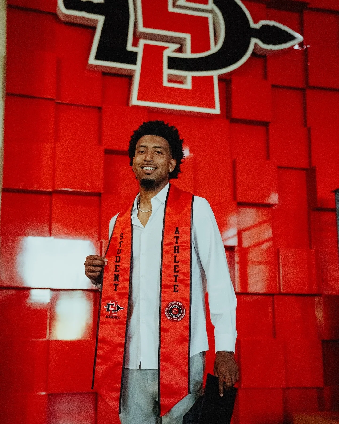 A young man dressed in a white shirt and beige pants, standing in front of a red wall with the San Diego State University logo, wearing a red stole that reads 'Student Athlete' and holding a graduation cap.