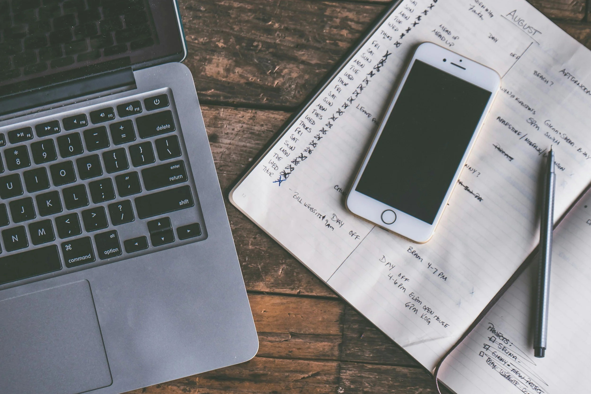 A workspace with a laptop, a smartphone, a pen, and a planner on a wooden table. The planner has handwritten notes and a checked calendar for August.