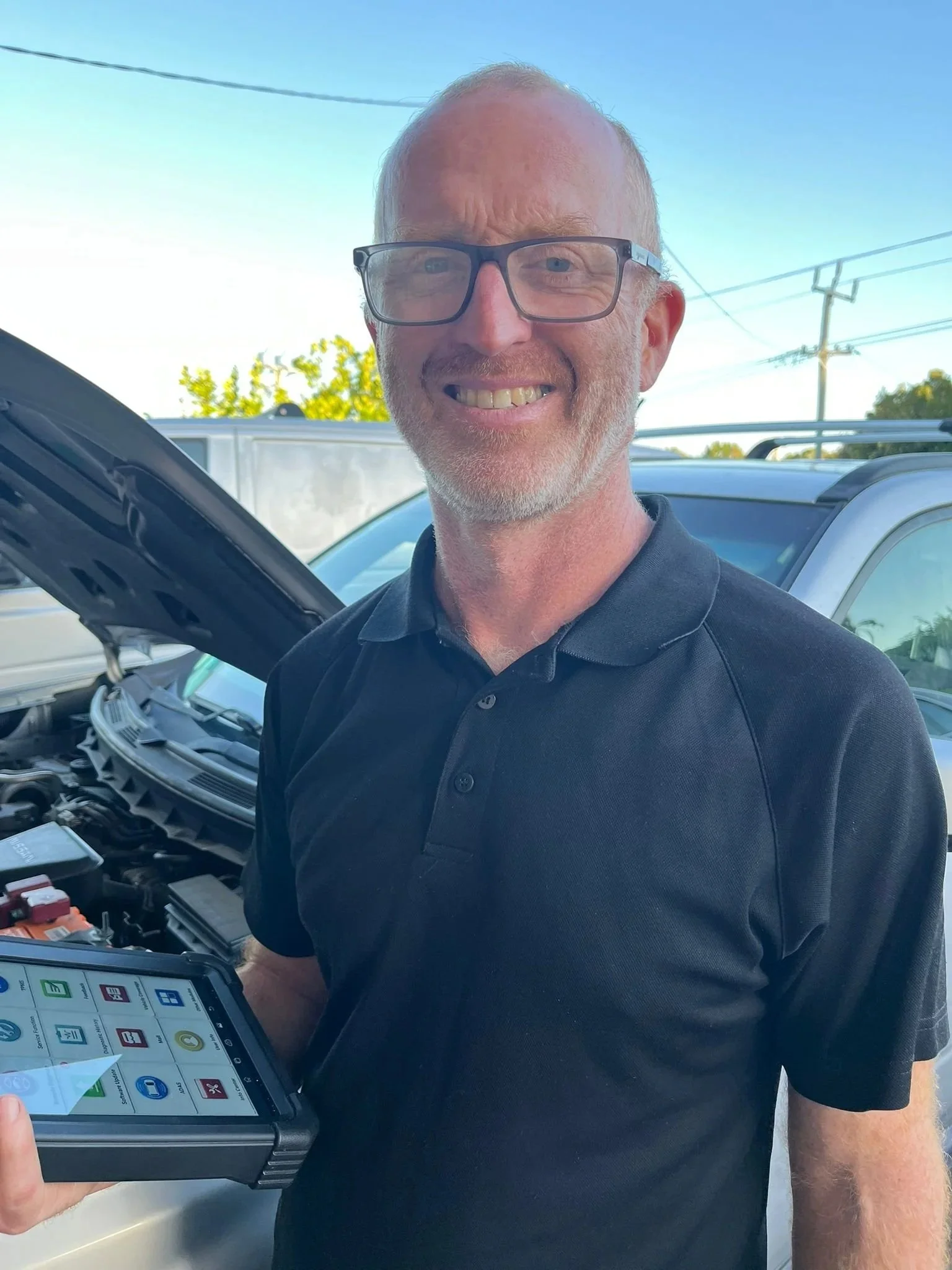 A man with glasses smiling and holding a tablet device stands in front of a white vehicle with its hood open, outdoors with other vehicles and power lines in the background.