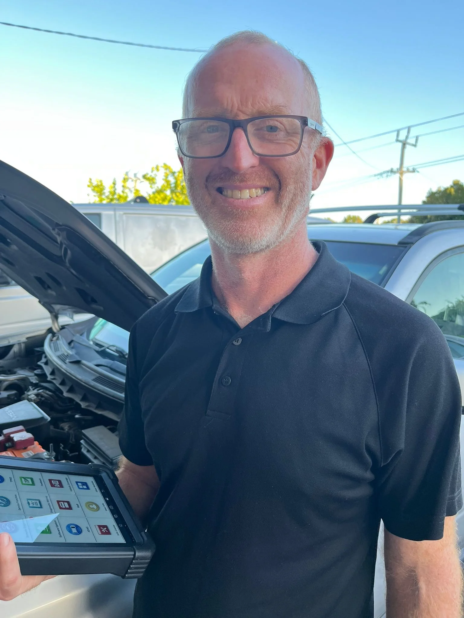 A man smiling outdoors next to a car with its hood open, holding a diagnostic tablet.