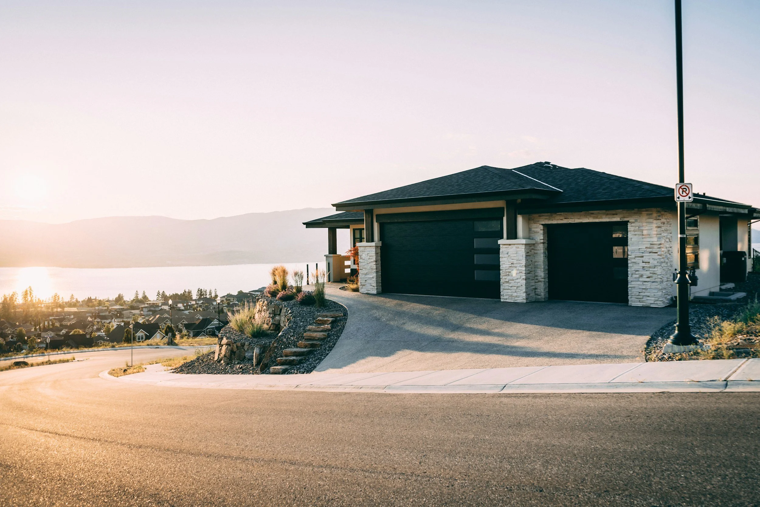 Modern house with a stone facade and two black garage doors on a sloped driveway, overlooking a lake and valley at sunset.
