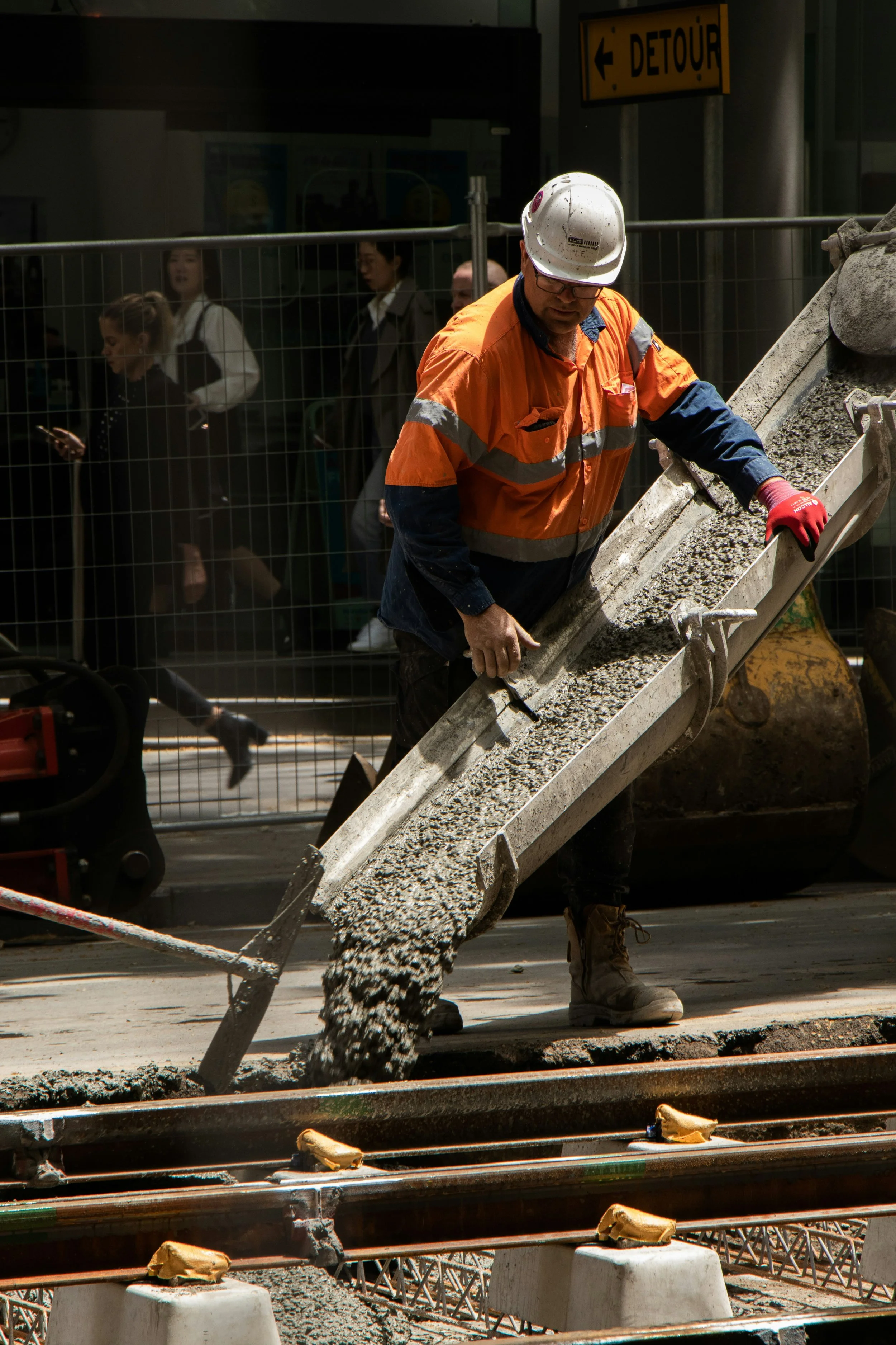 A construction worker pouring concrete on a sidewalk with a conveyor belt at a construction site, with people walking past in the background.