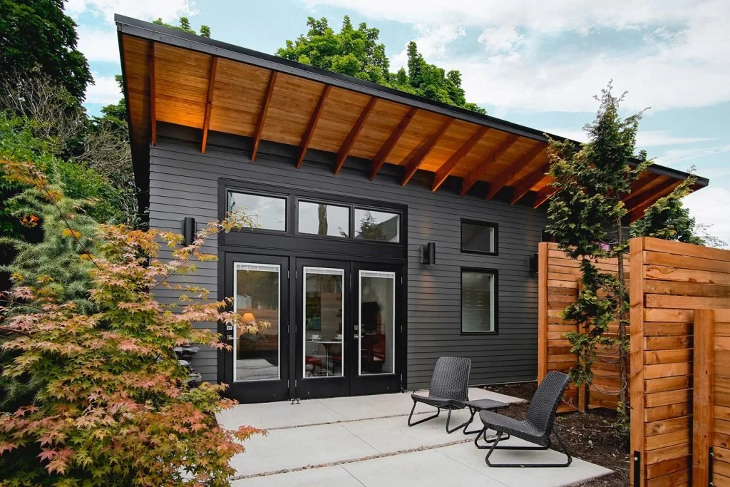 Modern black house with large glass doors and windows, wooden overhang roof, outdoor patio with two black chairs, surrounded by trees and wooden fence, during daytime.