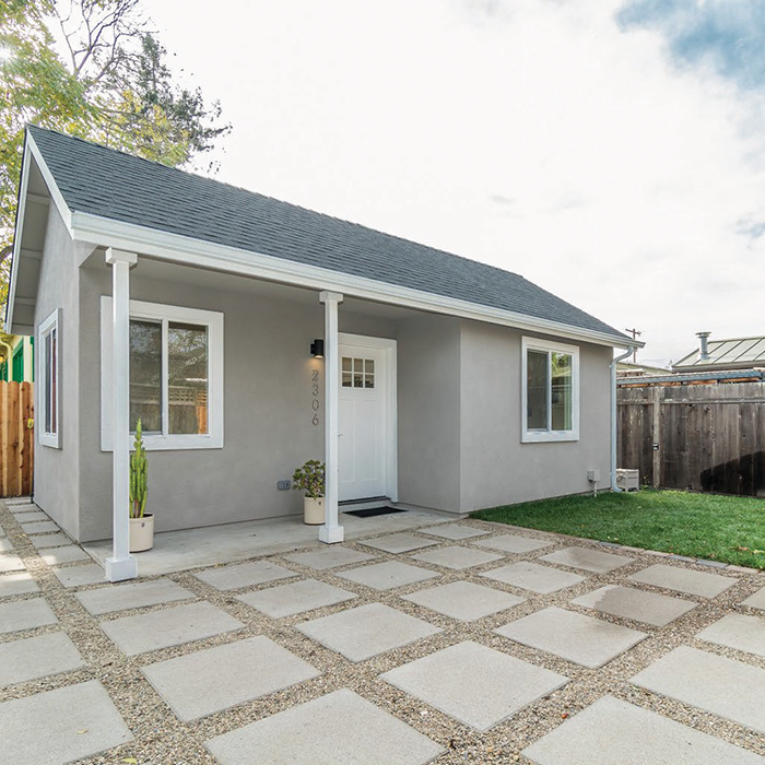 Front exterior of a small, modern house with a gray facade, white trim, and a pitched roof. The house has two large windows, a white door, and potted plants near the entrance. The driveway features large square concrete pavers with gravel in between.