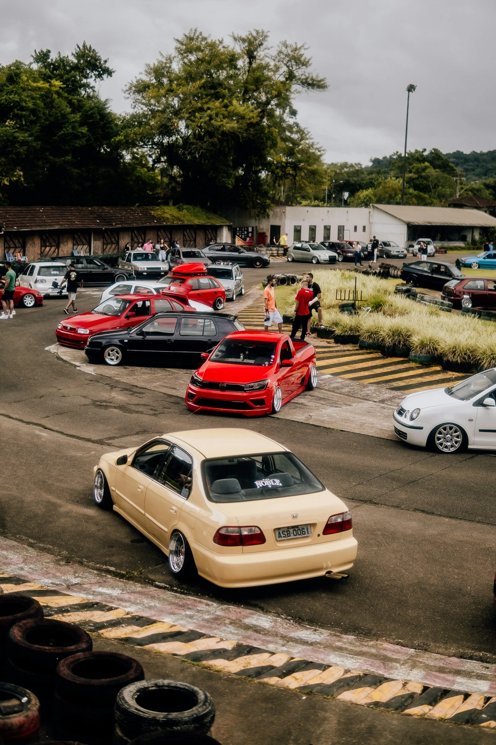 A car meet with various modified and stock cars, people socializing, and a cloudy sky in the background.