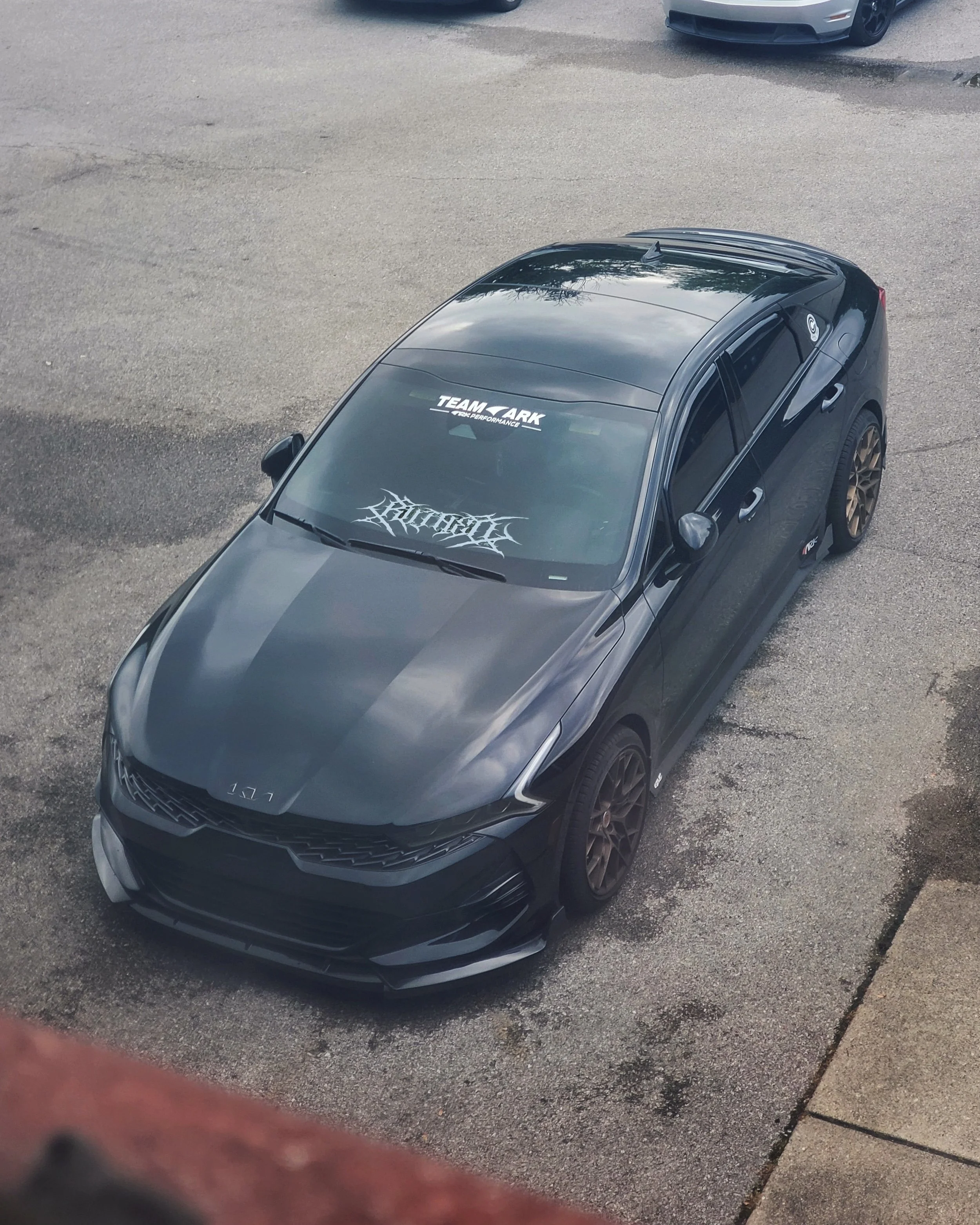 A black sports car parked on a rough asphalt surface, seen from above, with tinted windows and custom decals on the windshield and side windows.
