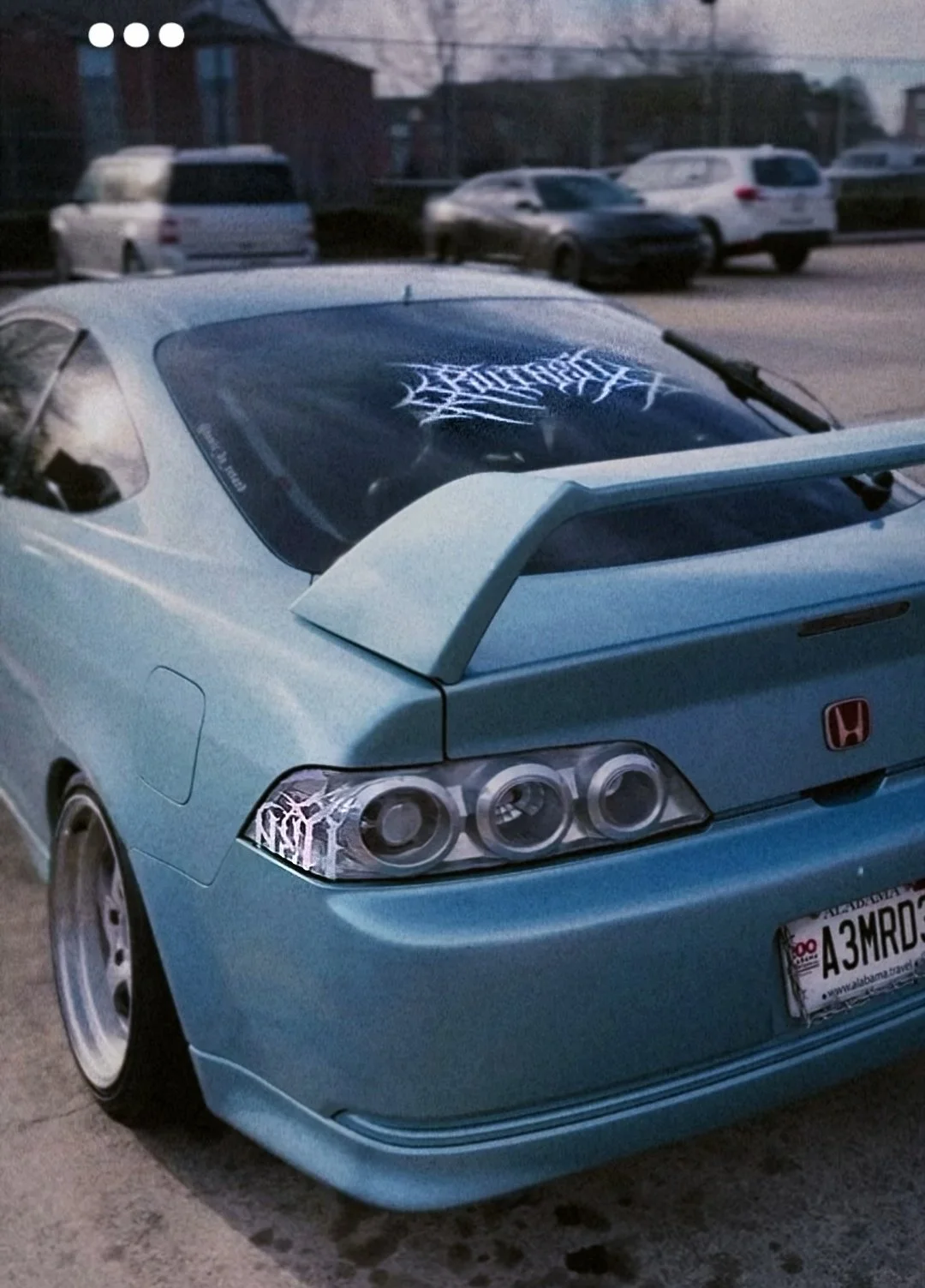 A blue Honda car with a large rear spoiler and custom tail lights, parked on the street at dusk, with a white sticker of an intricate, spiky design on the rear windshield.
