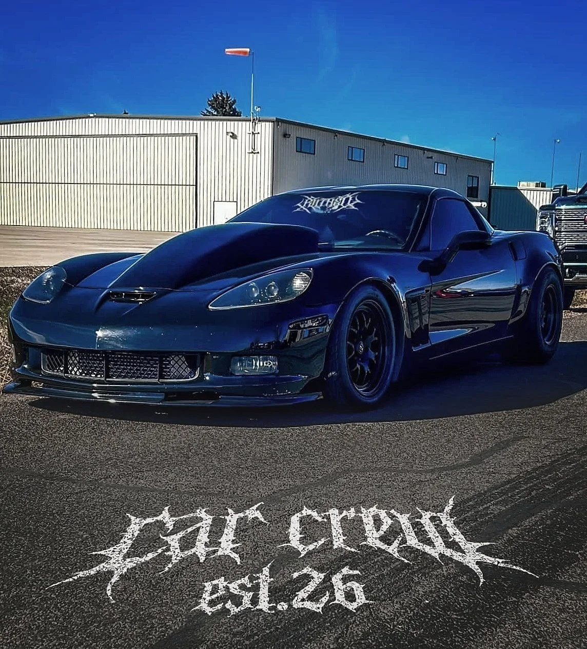 Black sports car with a vented hood and tinted windows parked on asphalt in front of a beige industrial building. Wind turbine blades are in the background under a clear blue sky. The words "car crew" and "EST. 26" are written in white gothic-style font on the road in front of the car.