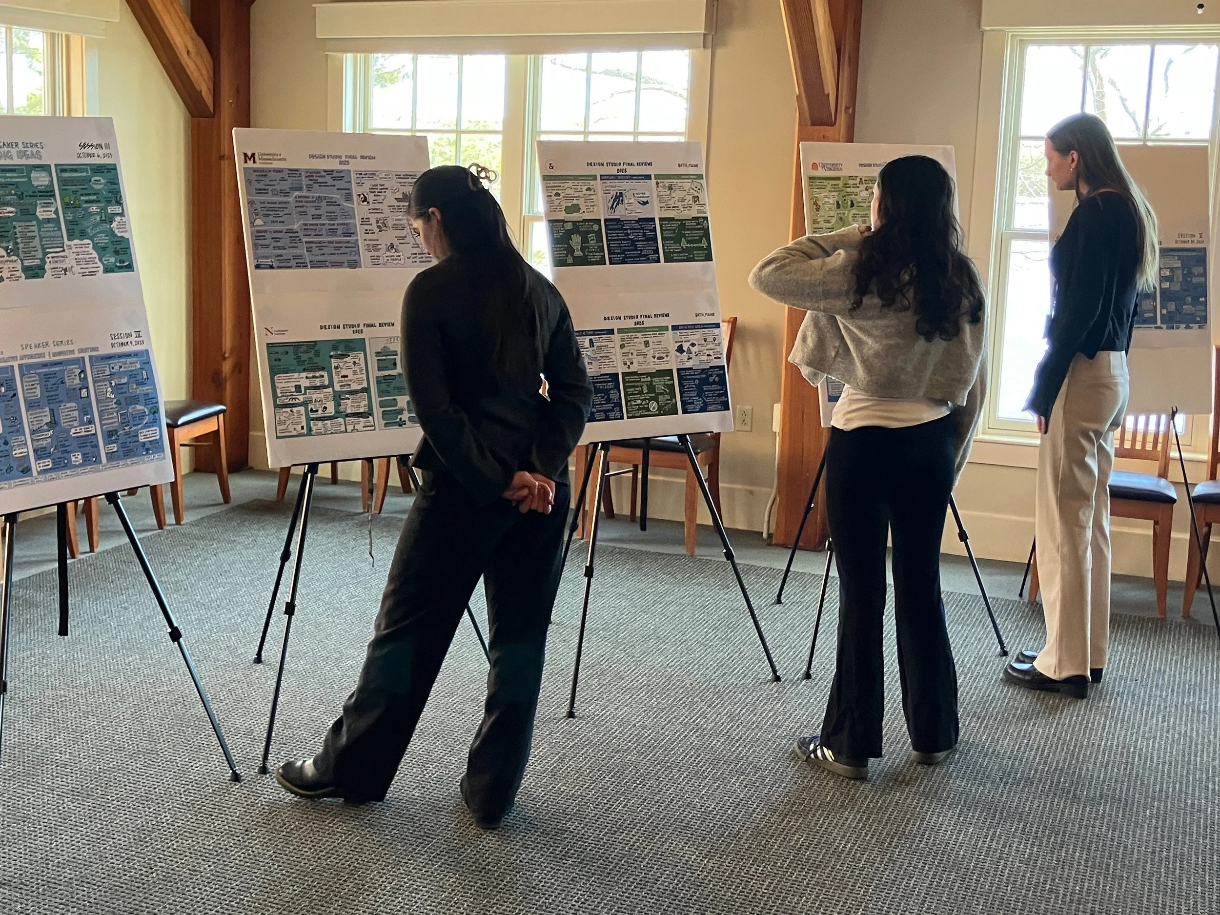 Three women stand in front of presentation boards inside a room with large windows. The women are engaged with the displays, which contain various infographics and text.