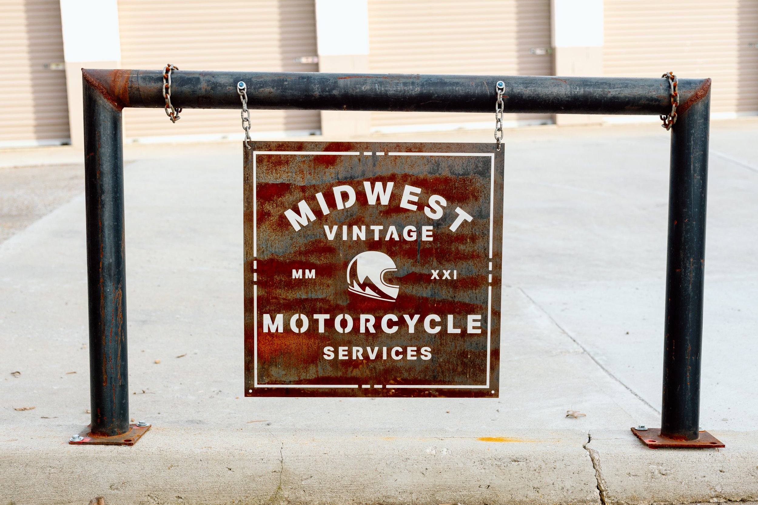 Rustic metal sign hanging from black metal frame advertising Midwest Vintage Motorcycle Services, with a wave logo in the center, set outdoors on a concrete sidewalk.