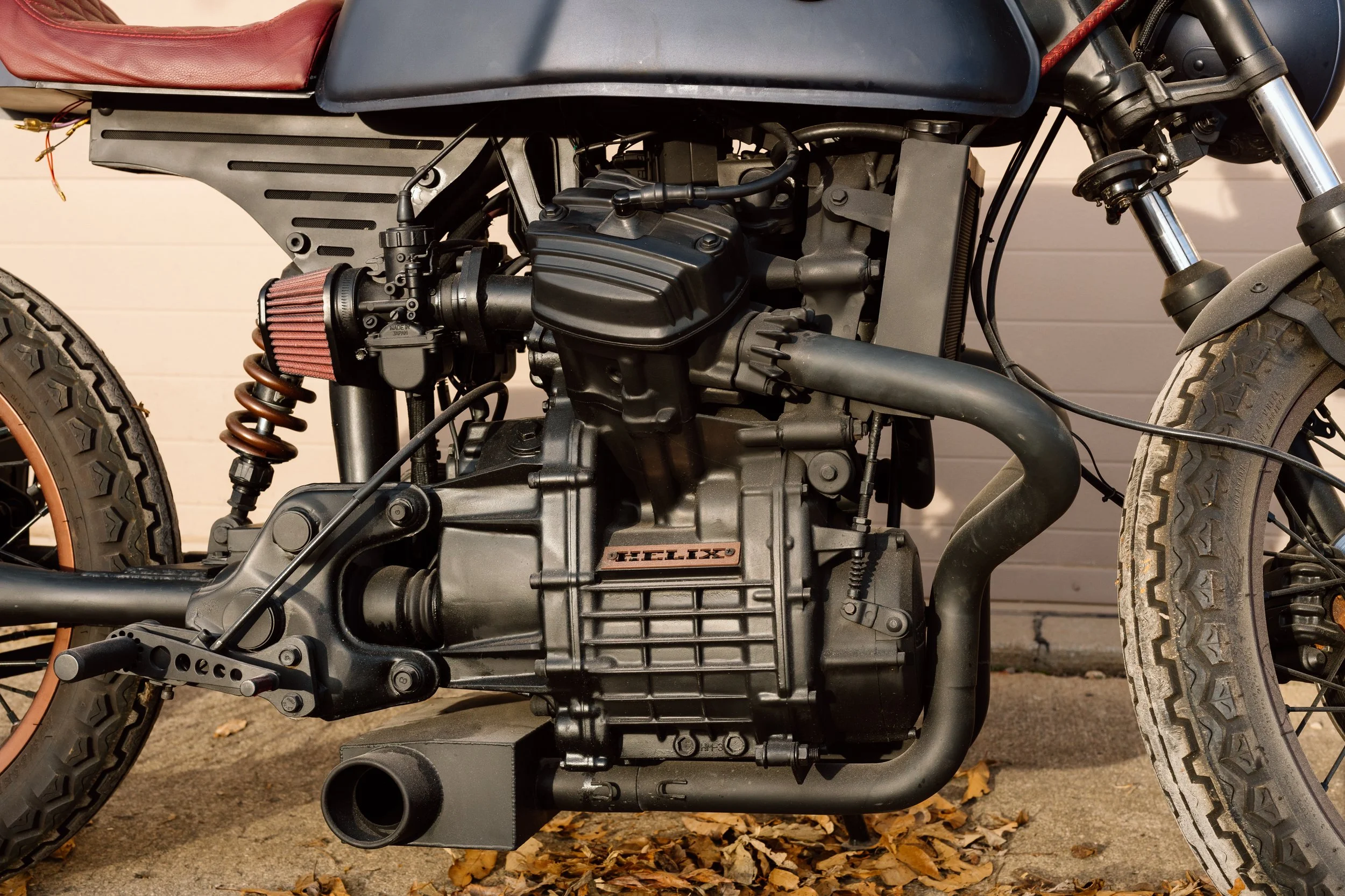 Close-up of a motorcycle engine with black frame and tires, surrounded by fallen leaves on ground.
