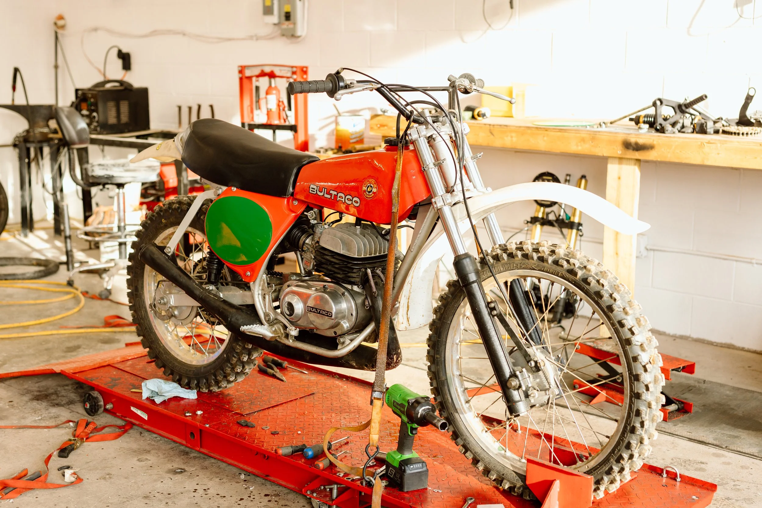 Vintage Bultaco dirt bike on a red repair stand in a workshop, surrounded by tools and equipment.