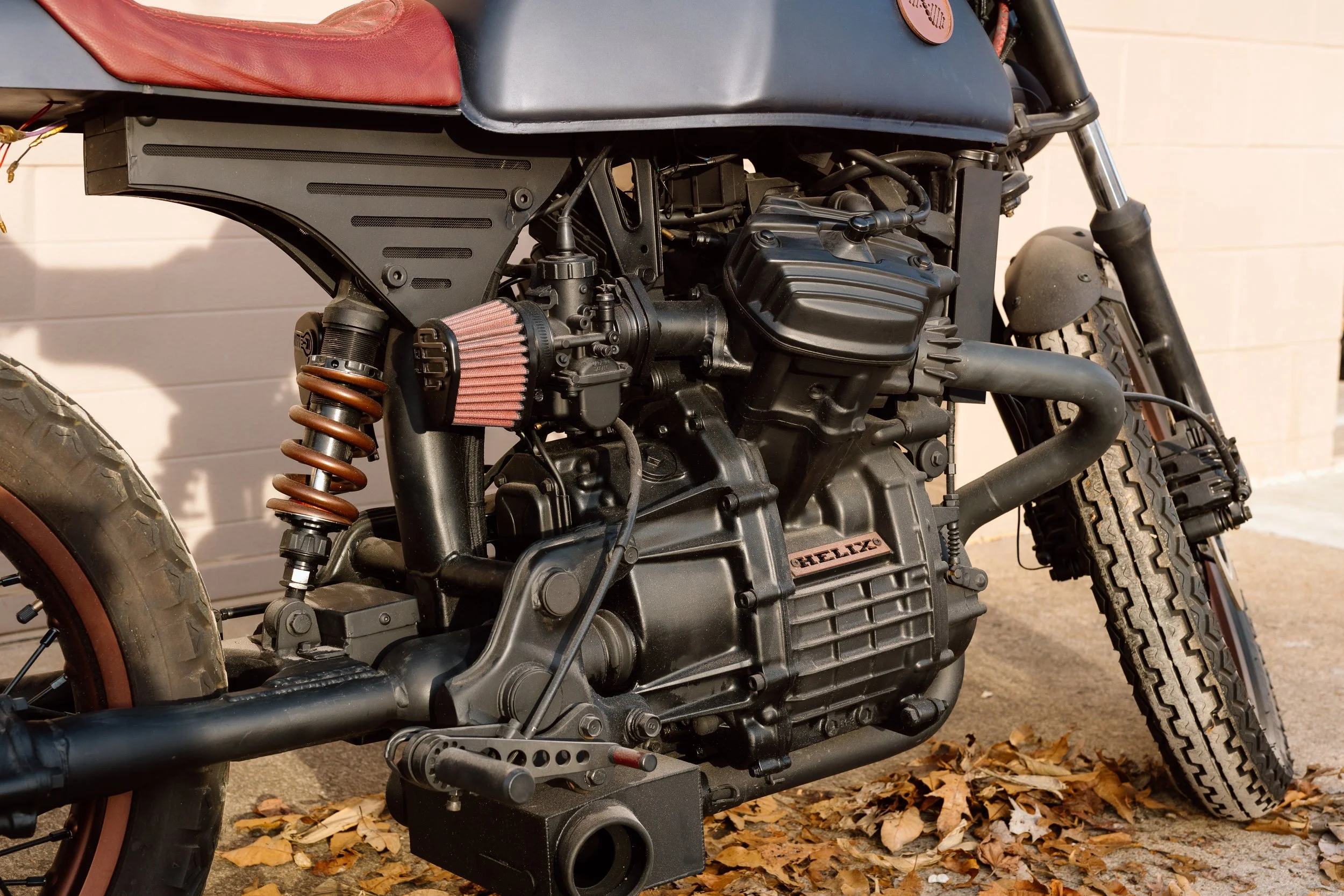 Close-up of a black motorcycle engine and suspension with a red air filter, set against a brick wall and fallen autumn leaves.