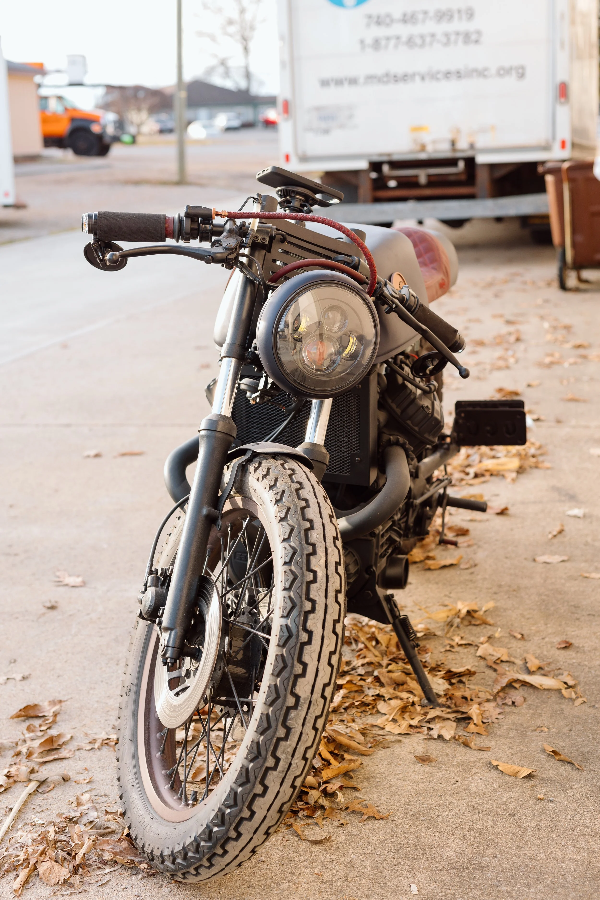 A vintage-style motorcycle parked on a sidewalk with fallen autumn leaves, a truck and some buildings in the background.