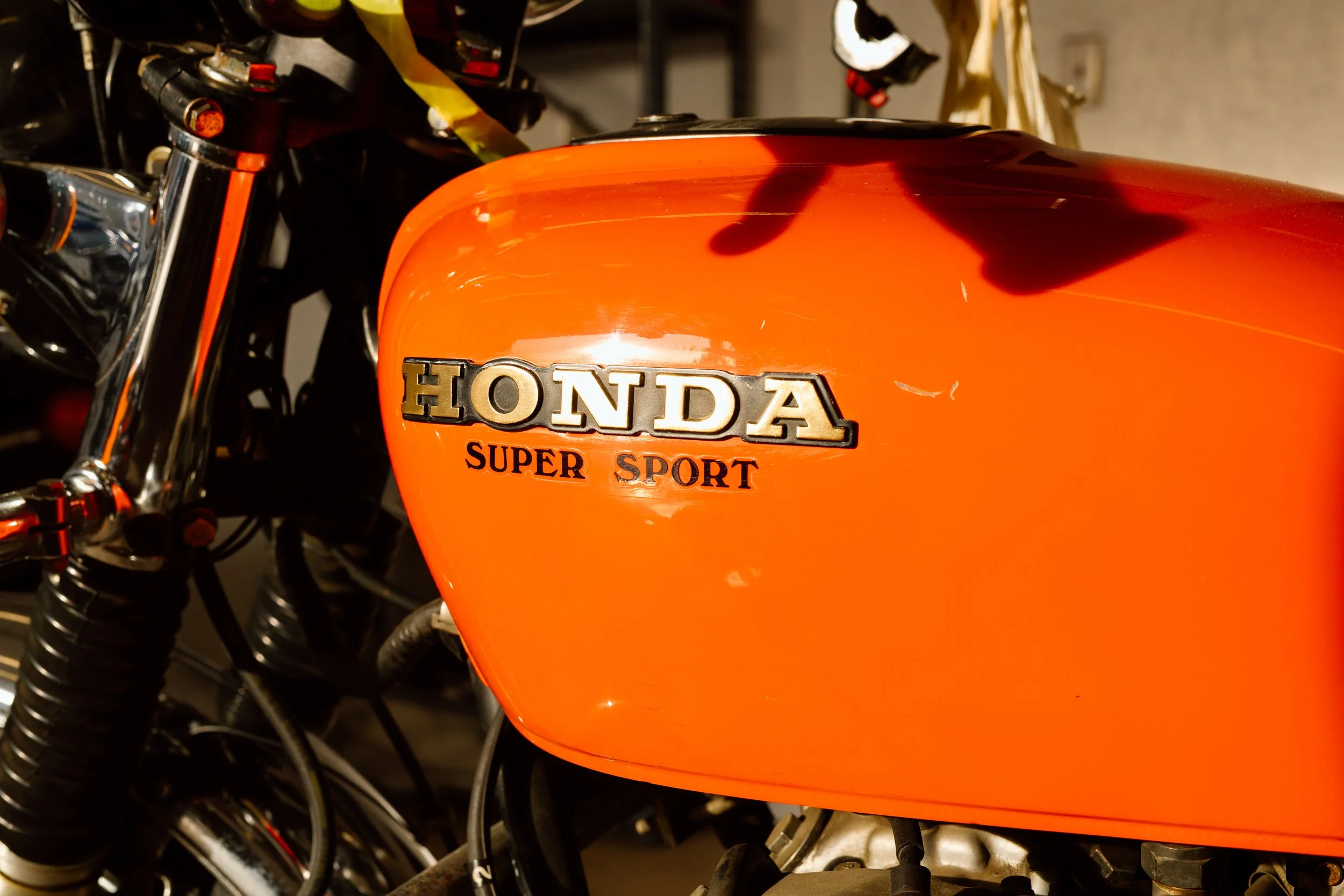 Close-up of an orange Honda Super Sport motorcycle fuel tank with chrome emblem, showing motorcycle handlebar and mechanical parts in the background.