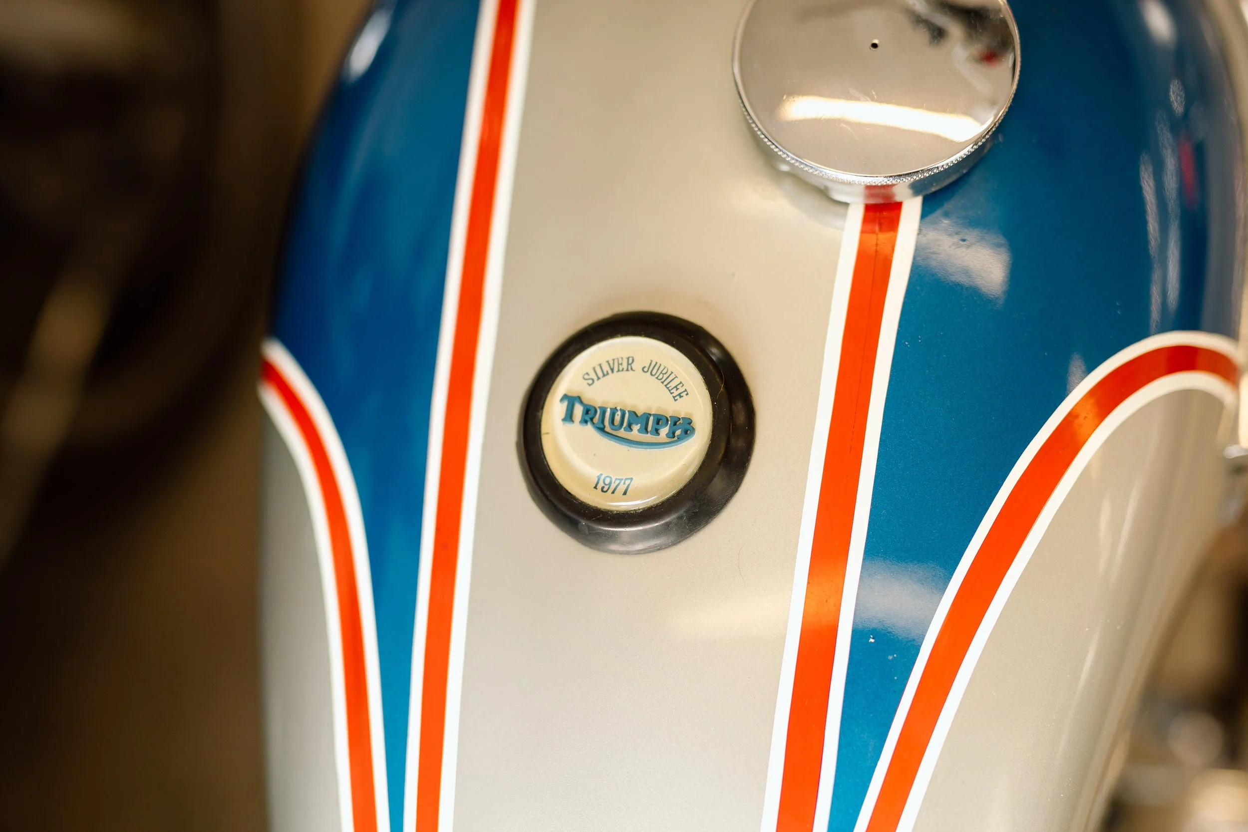 Close-up of a vintage Triumph motorcycle fuel tank with a badge reading 'Silver Jubilee Triumph 1977' and red, white, and blue piping.