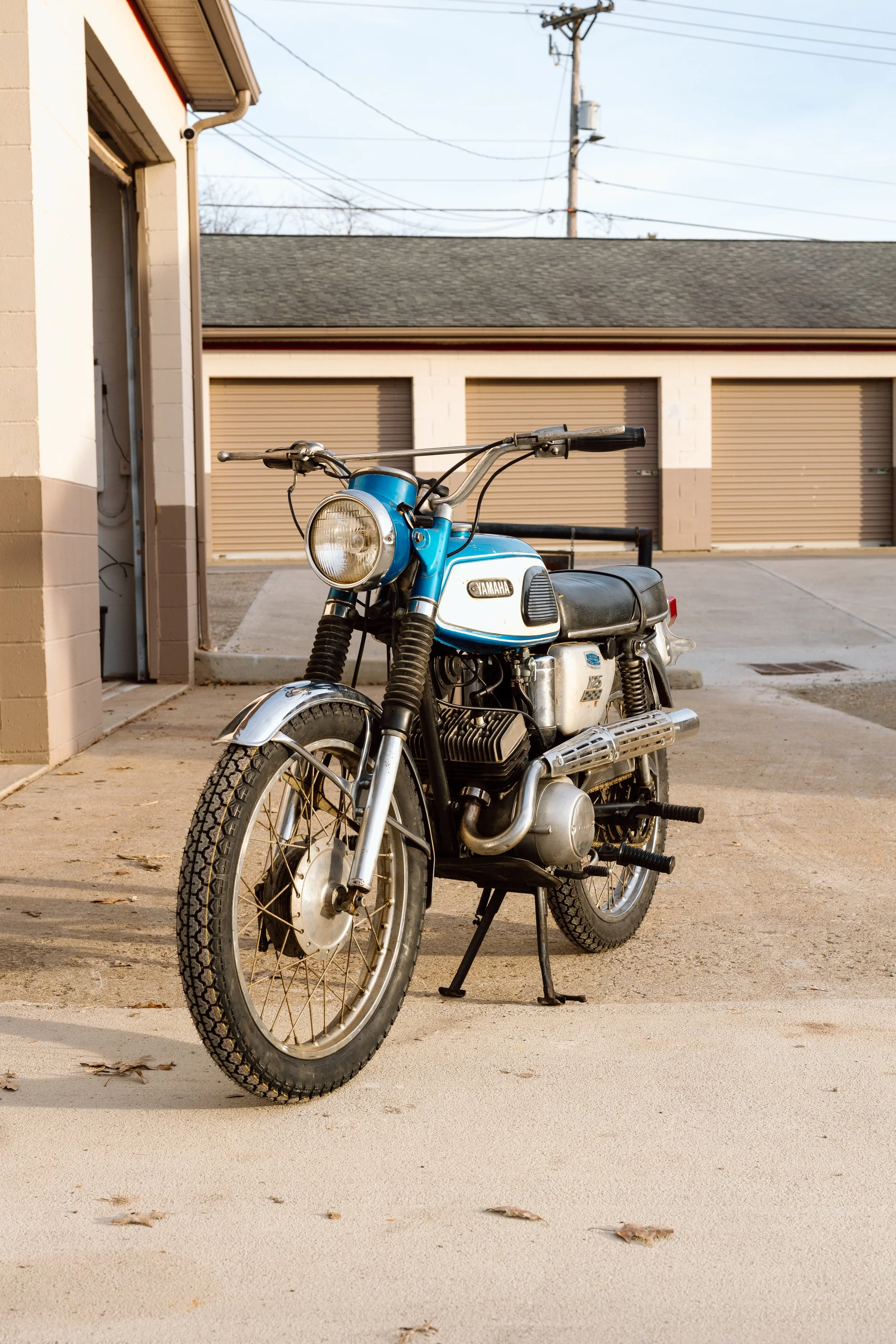 A vintage blue and silver Yamaha motorcycle parked outside a garage on a concrete driveway, with closed garage doors in the background.