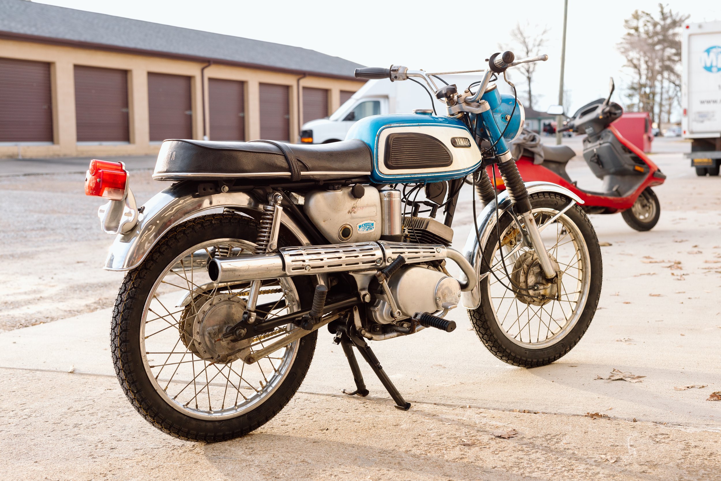 A vintage Yamaha motorcycle parked on a sidewalk with a red scooter and parked trucks in the background.
