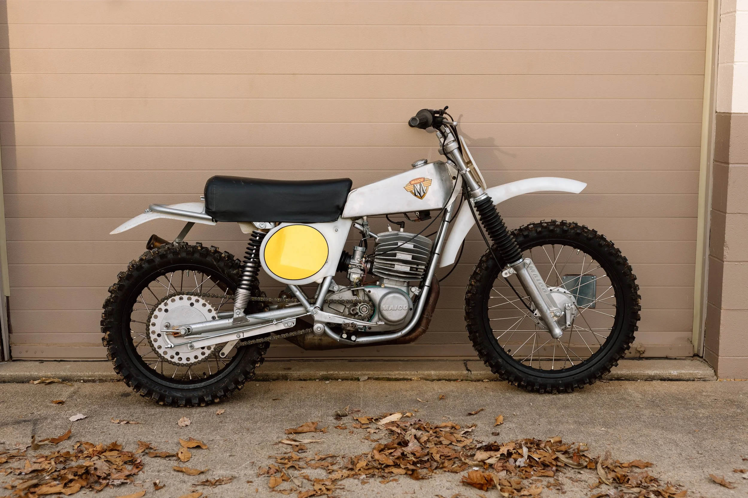 A vintage off-road motorcycle with knobby tires and a black seat, parked against a beige garage door with fallen leaves on the ground.