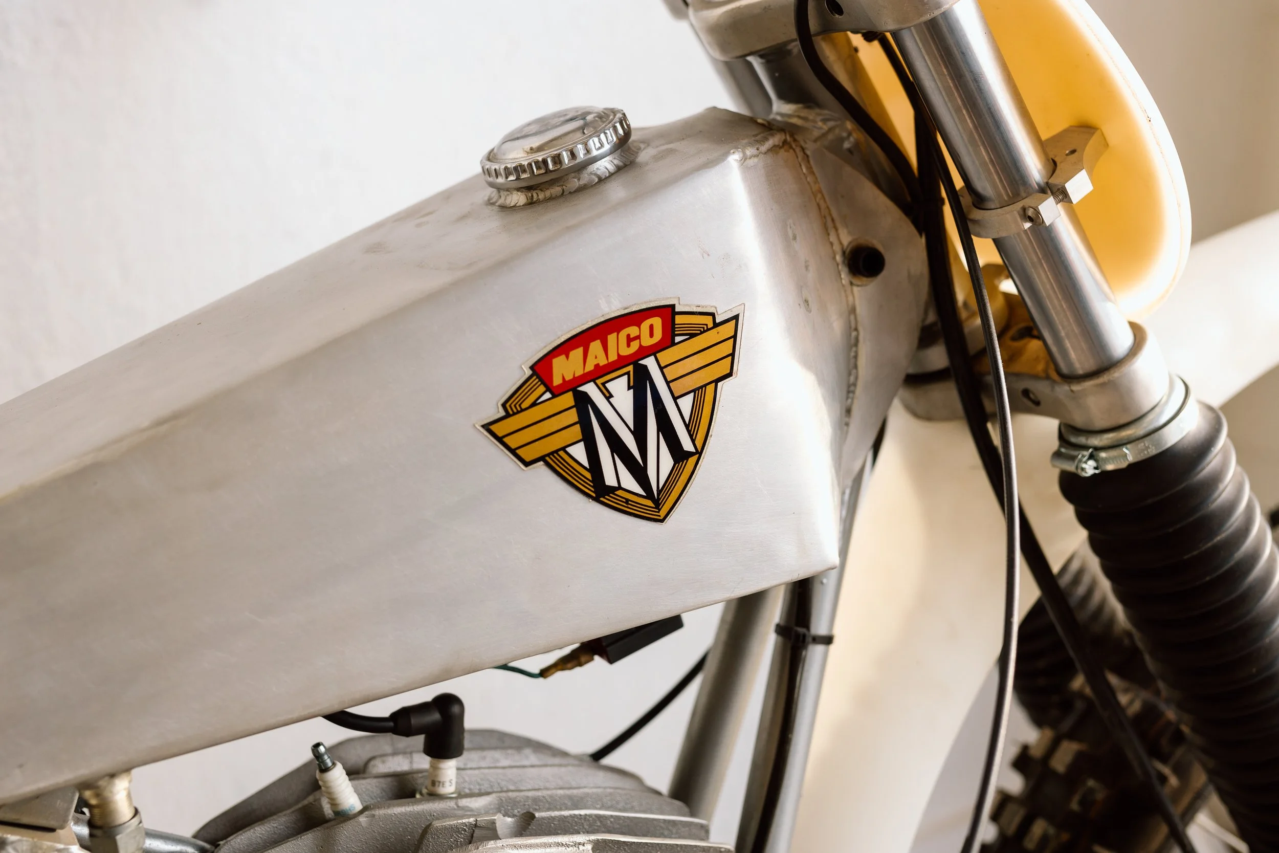 Close-up of a vintage Maico motorcycle fuel tank with a Maico logo, showing the tank's metallic surface and part of the front fork and suspension.