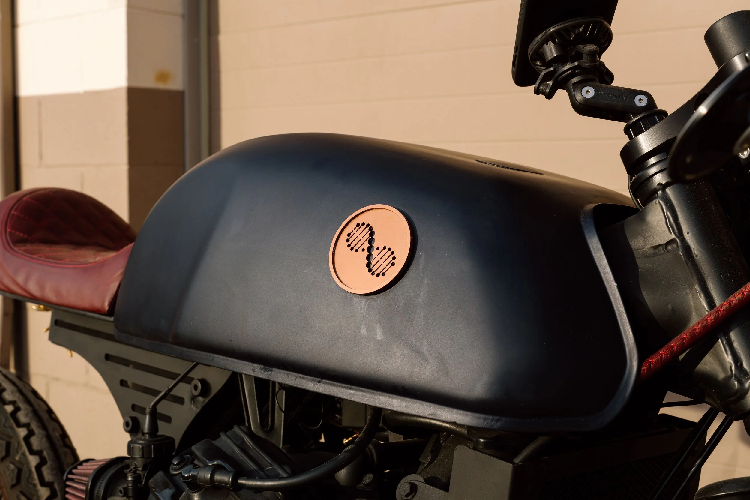Close-up of a black motorcycle fuel tank with a pink circular emblem featuring a stylized butterfly design, and part of a pink leather seat visible on the left.