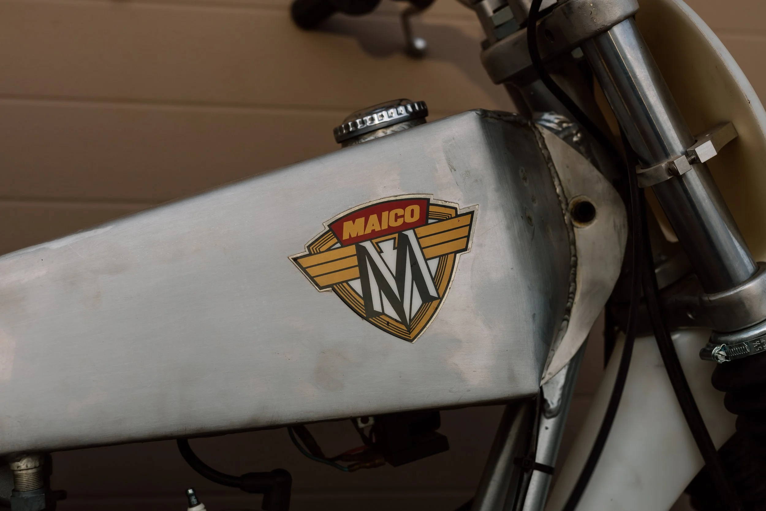 Close-up of a vintage Maico motorcycle fuel tank with an old Maico logo sticker, showing metal body, front fork, and wiring.