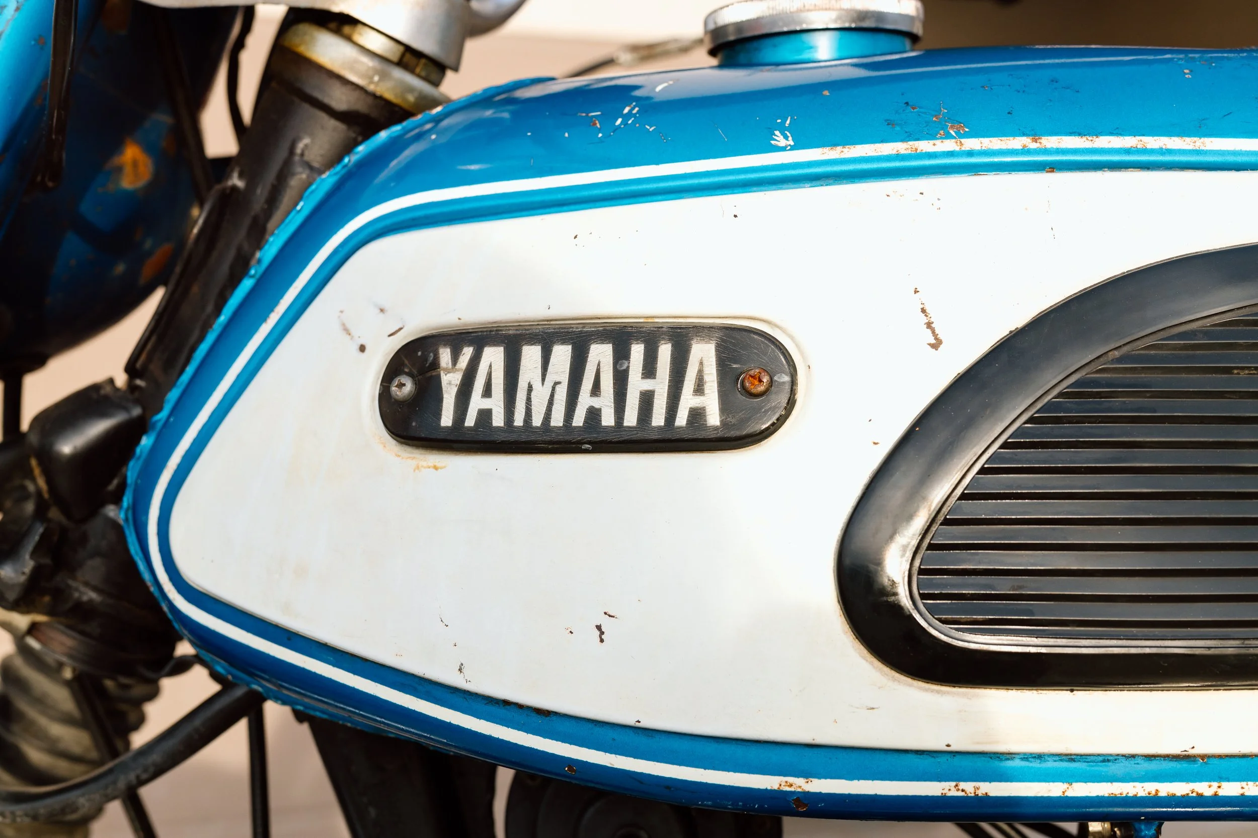 Close-up of a vintage Yamaha motorcycle fuel tank with blue and white paint, showing signs of rust and wear, and a black Yamaha emblem.