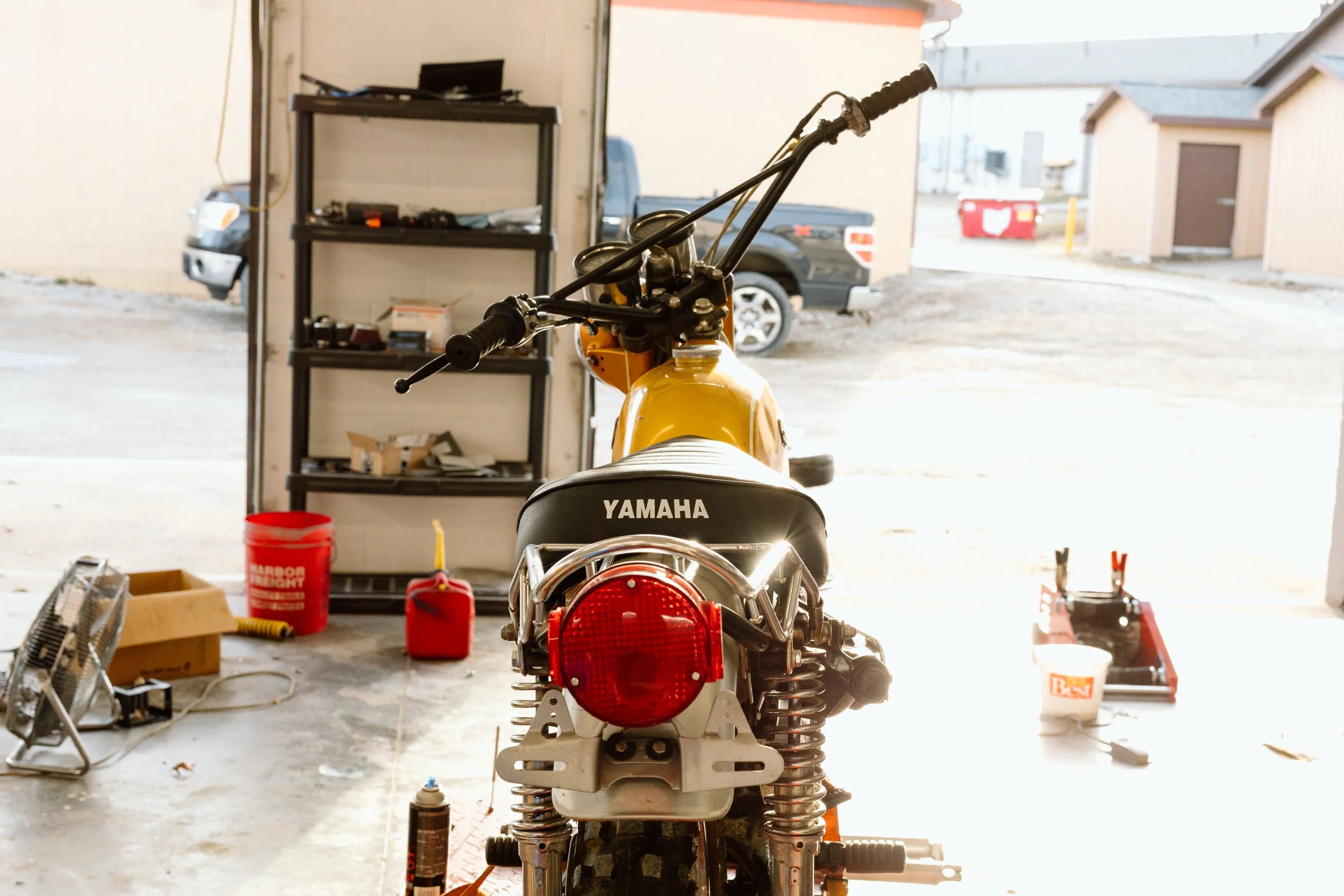 Rear view of a yellow Yamaha motorcycle in a garage, with tools and equipment nearby, and vehicles visible outside through the open garage door.